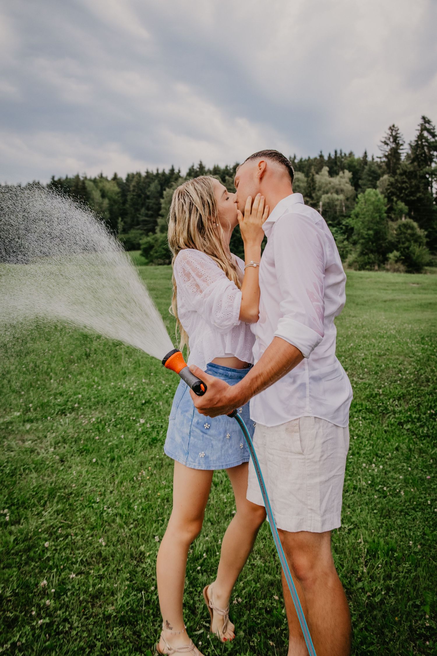 A couple kisses outdoors on green grass while holding a garden hose spraying water, with trees in the background.