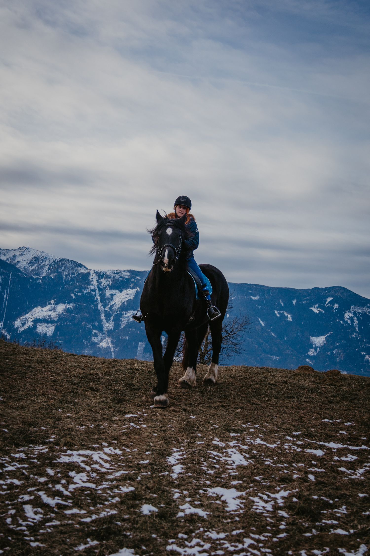 Two horses and their riders traverse a snowy mountain trail with dramatic mountain peaks in the background.