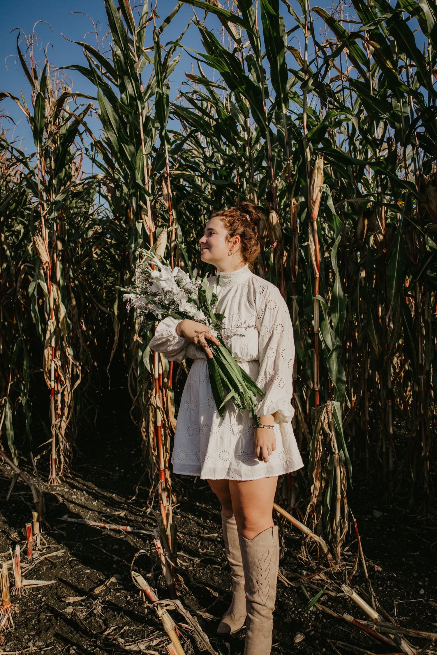 A figure in a white eyelet dress poses with flowers in a cornfield at golden hour.