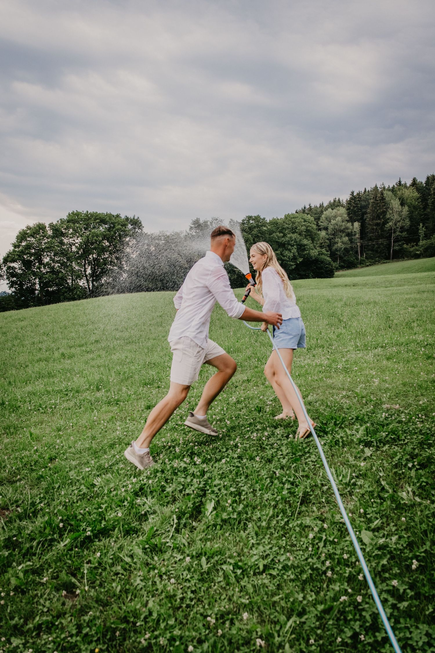 Couple balancing together on a slackline in a lush green meadow with trees and cloudy sky in background.