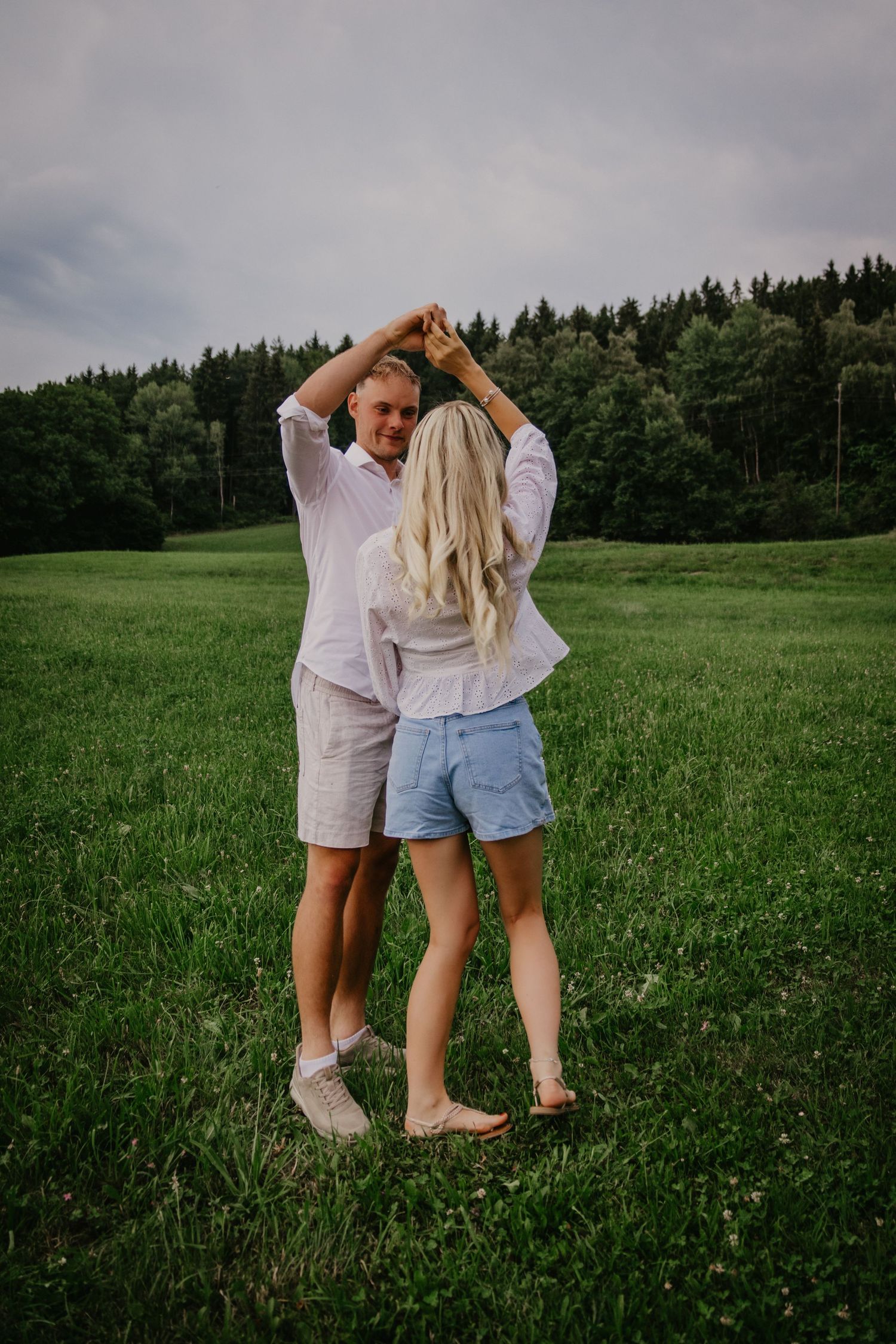 Couple dancing together in white outfits on a lush green field with forest backdrop on a cloudy day.