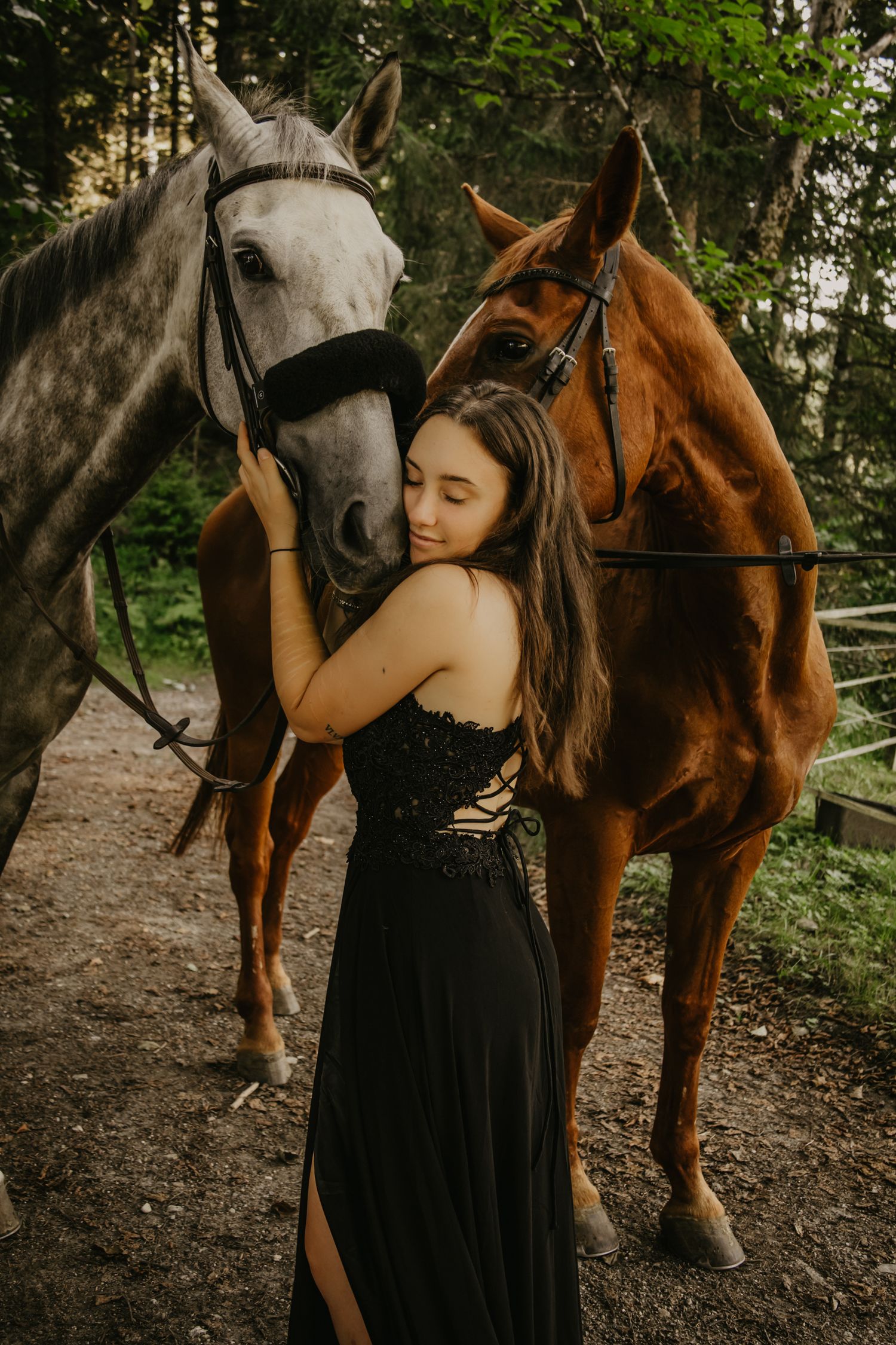 Two horses, one grey and one bay, stand with someone in a black dress on a forest path.