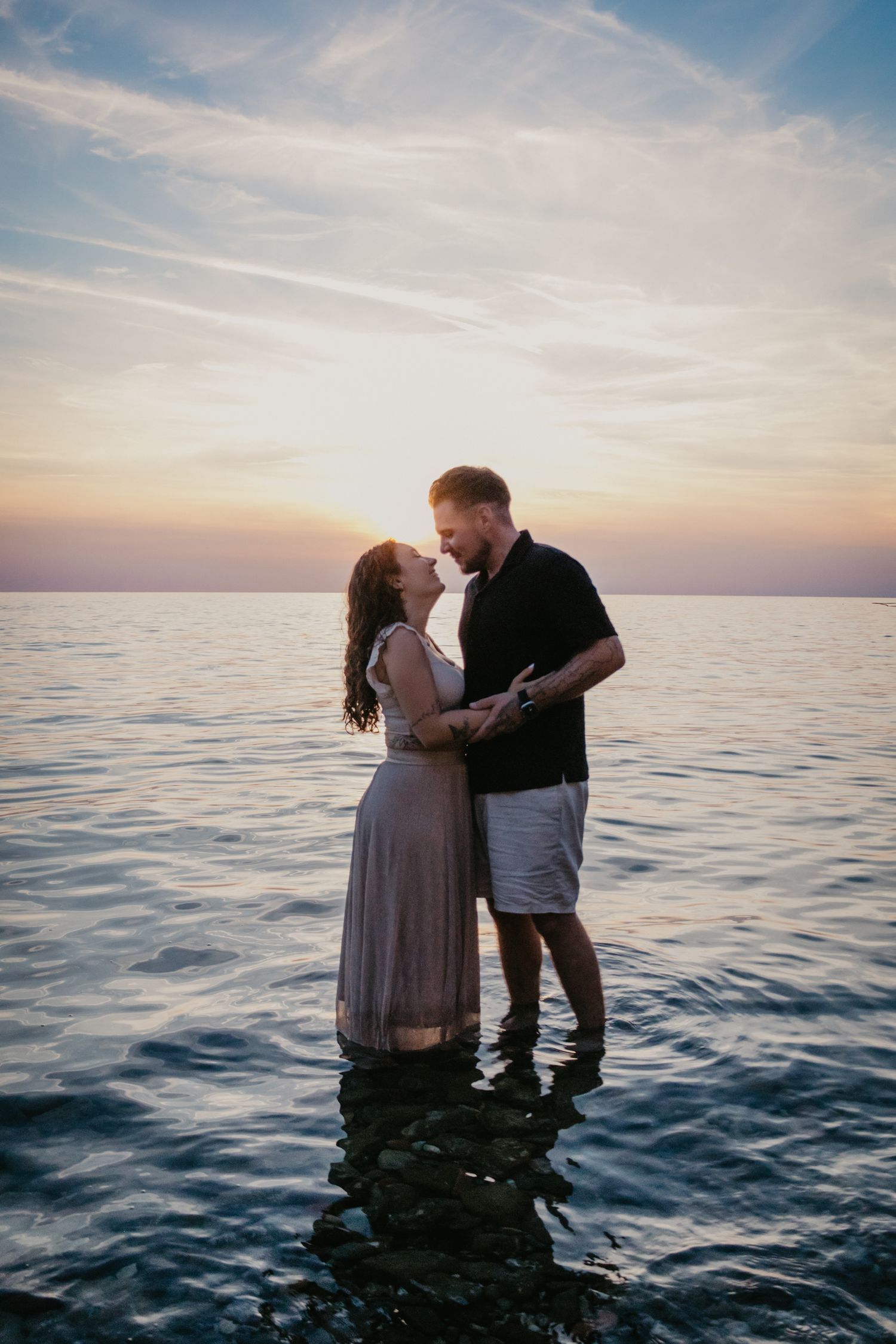 Two people share an intimate moment while standing in ocean water during sunset.