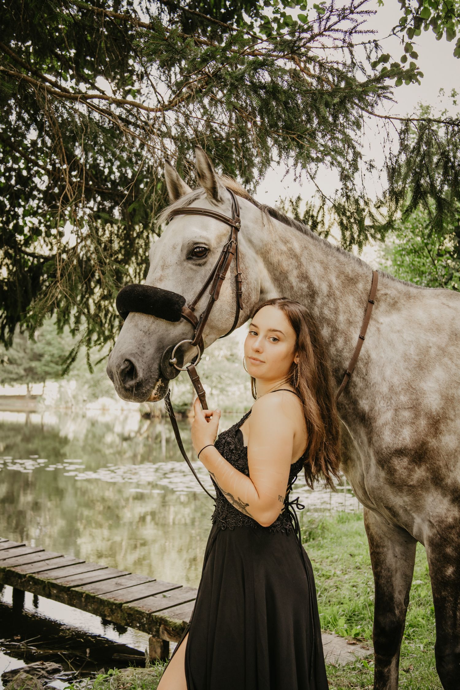 A dappled grey horse poses with its rider in a black formal dress by a wooden dock.