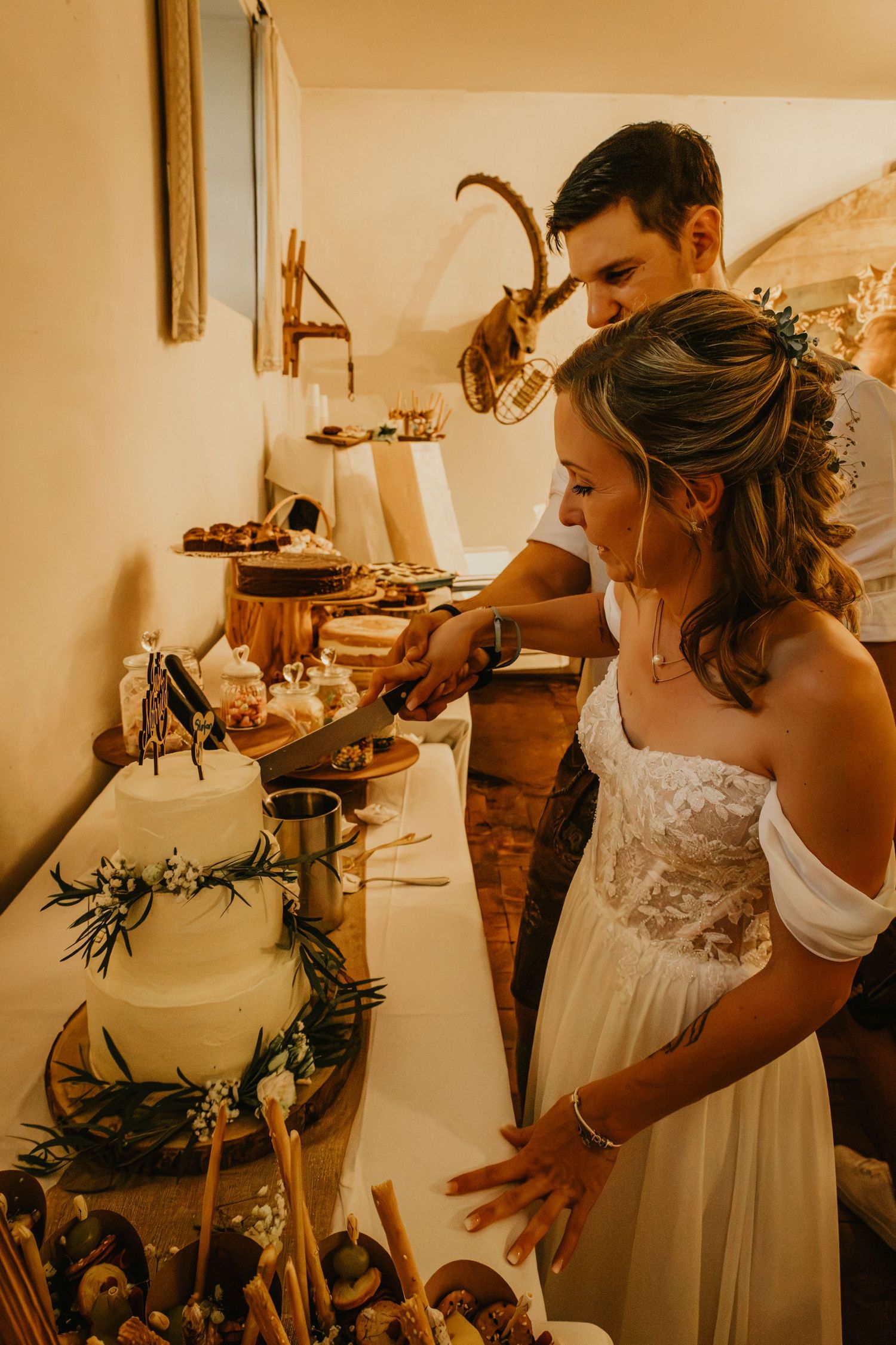 Bride and groom cutting their wedding cake together at a rustic reception table decorated with greenery.