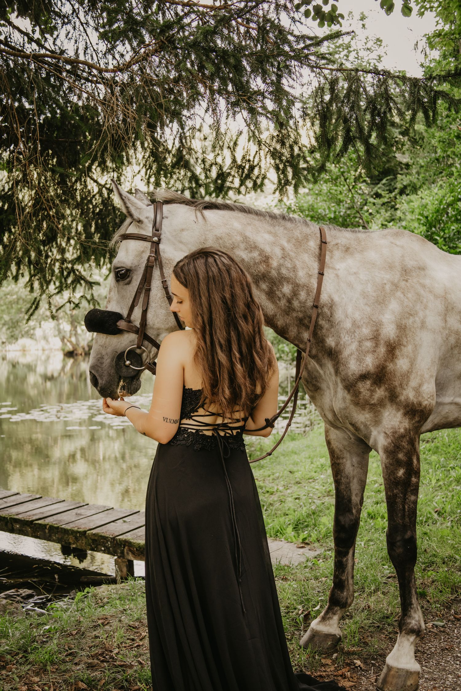 A grey horse stands beside someone in a black dress near a pond under overhanging tree branches.