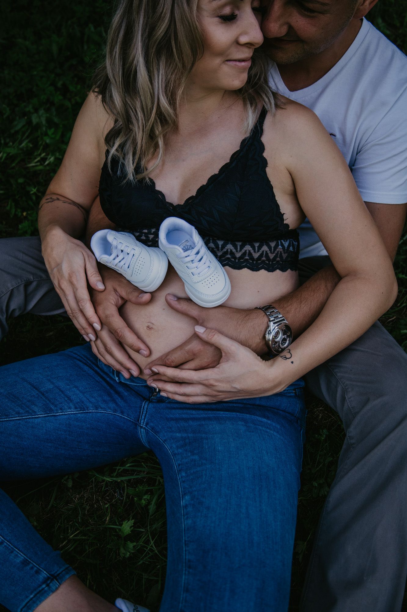 A couple holds baby shoes while embracing outdoors.