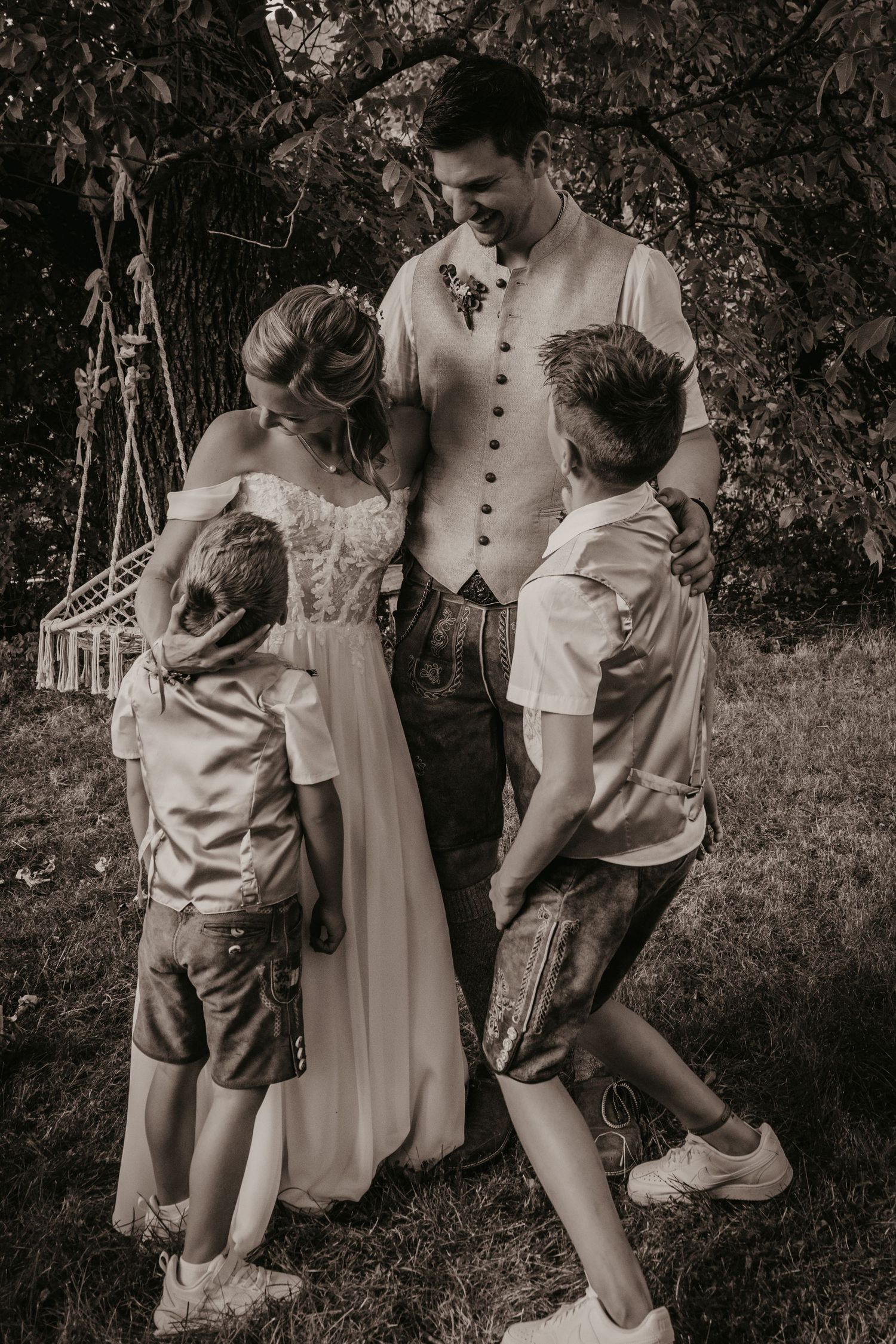 Black and white photo of a bride and groom in Bavarian attire embracing two boys outdoors under a tree.