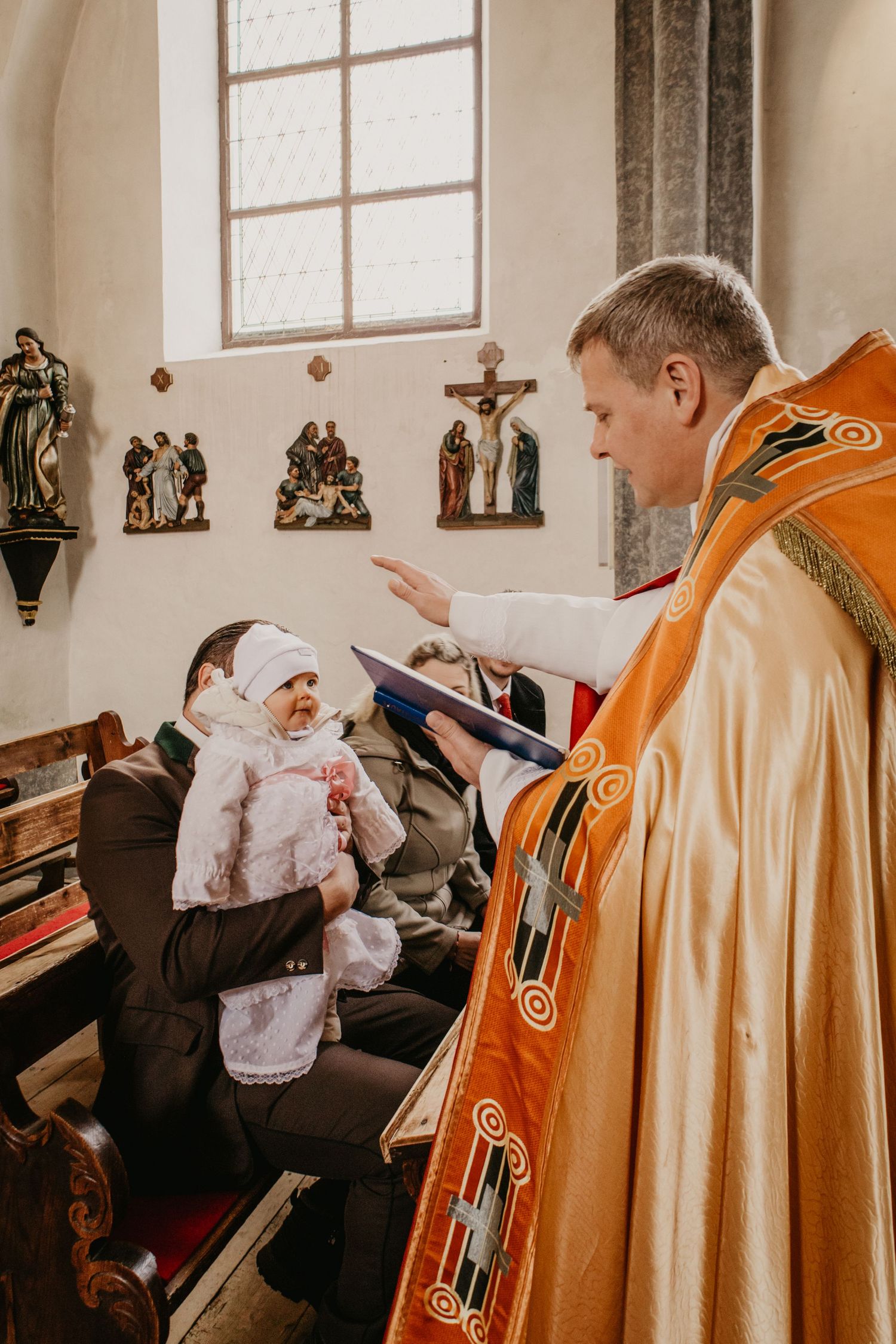 A priest in ornate golden vestments blesses a baby in white christening attire during a baptism ceremony in a church.