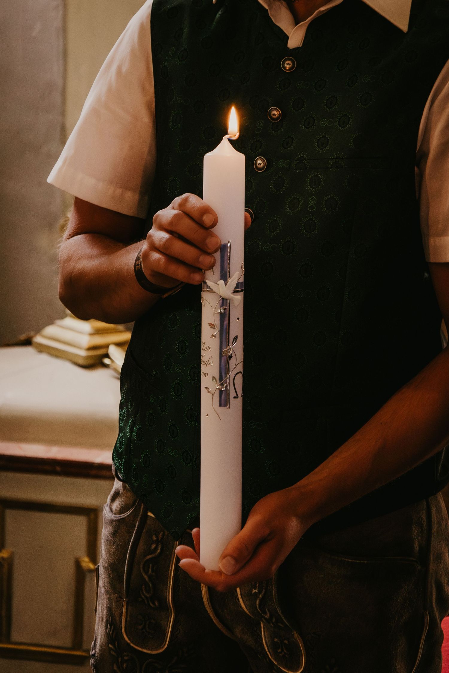 Person in a dark green vest and traditional leather trousers holds a lit white baptism candle with blue decorations.