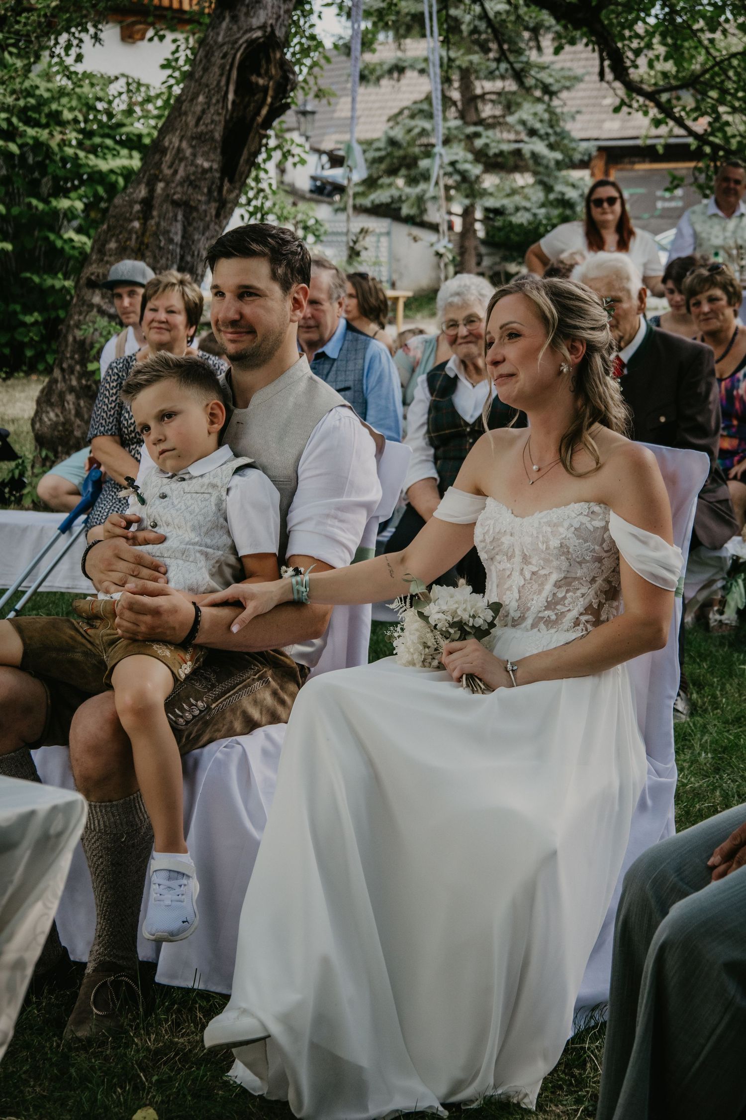 Bride and groom seated outdoors at wedding ceremony with young boy on grooms lap holding bouquet.