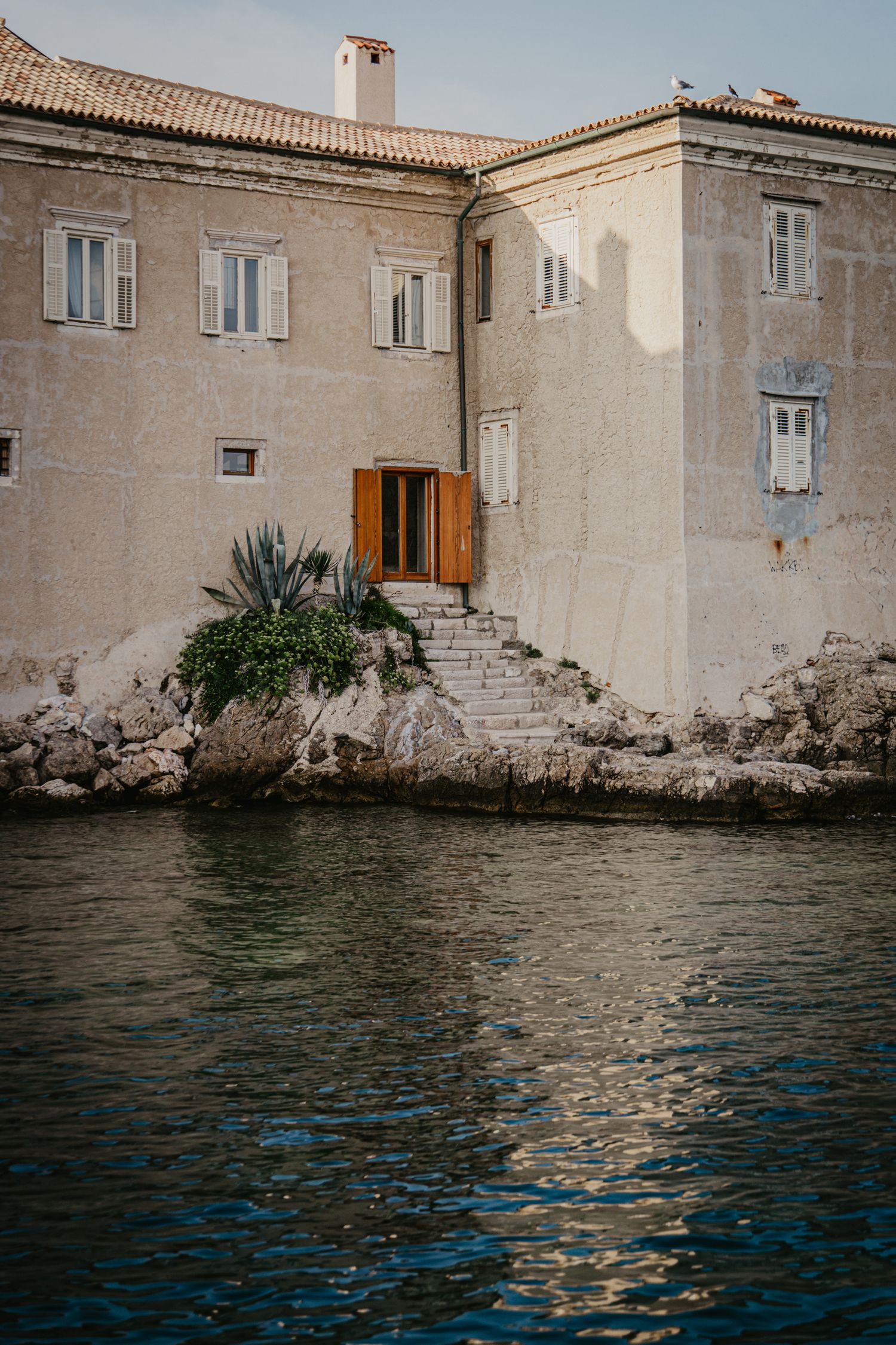 Historic waterfront buildings with stone walls reflecting in calm Mediterranean waters.