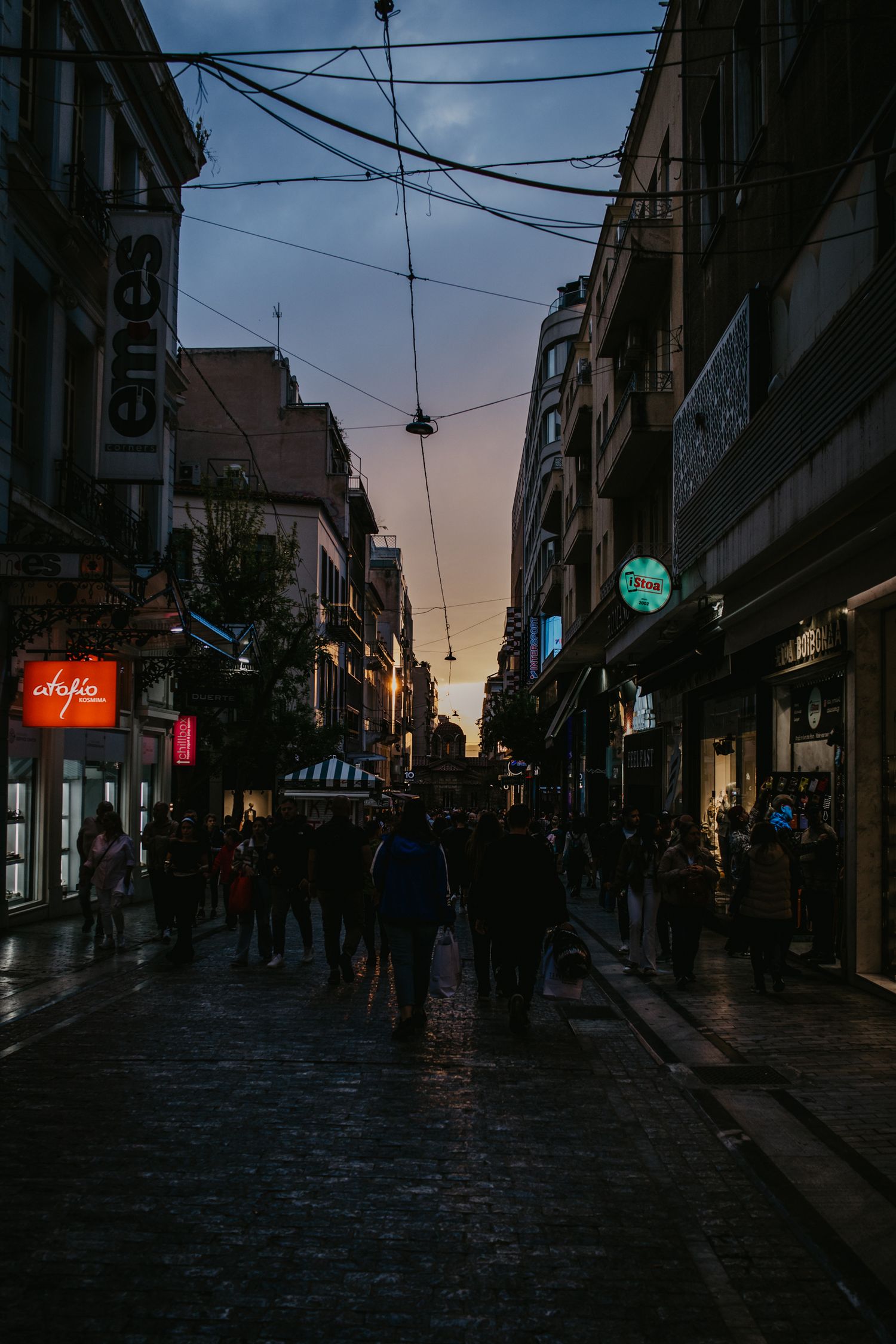 Dimly lit cobblestone street at dusk with neon signs and silhouettes.