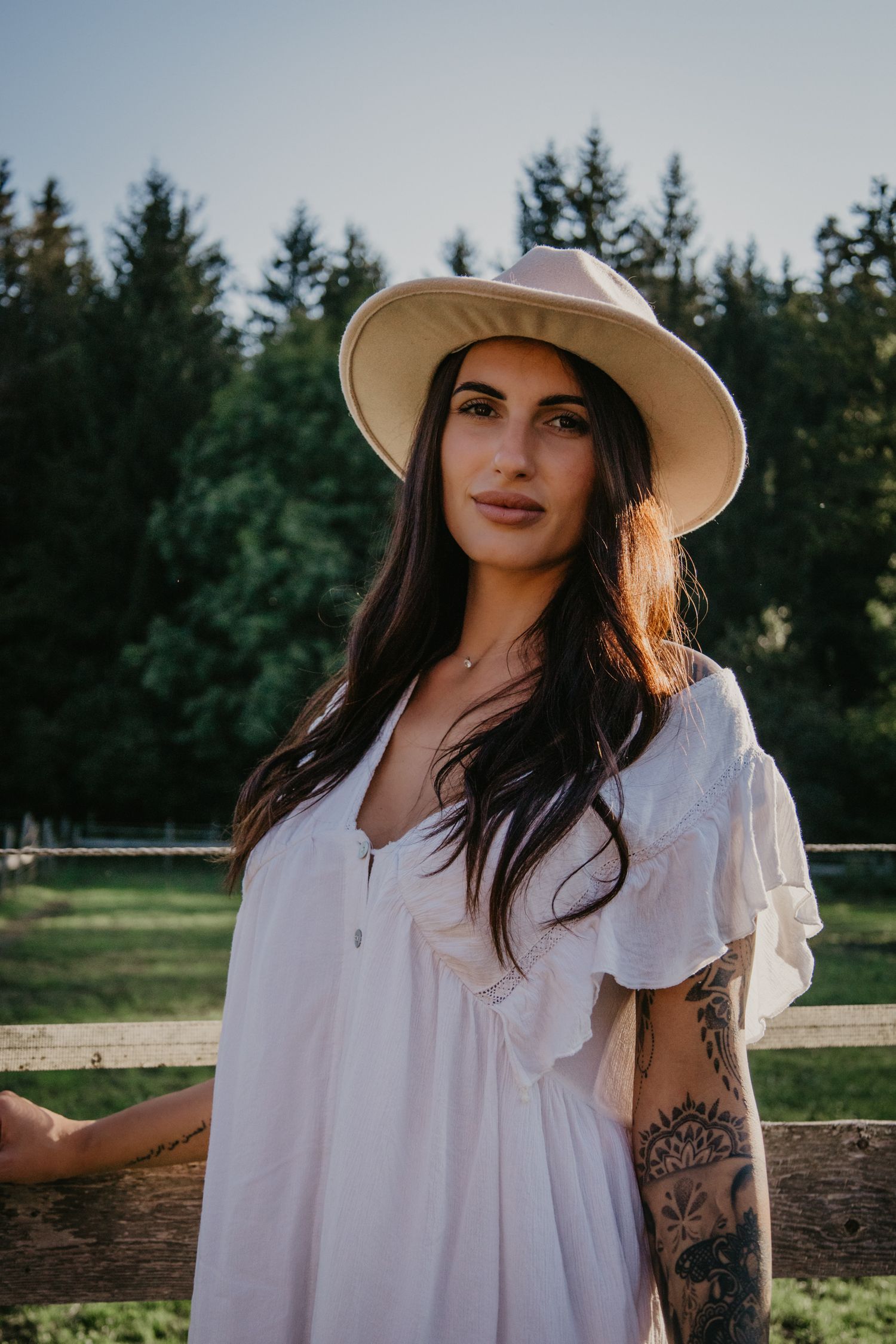 Countryside portrait in white dress and wide-brim hat against evergreen trees.