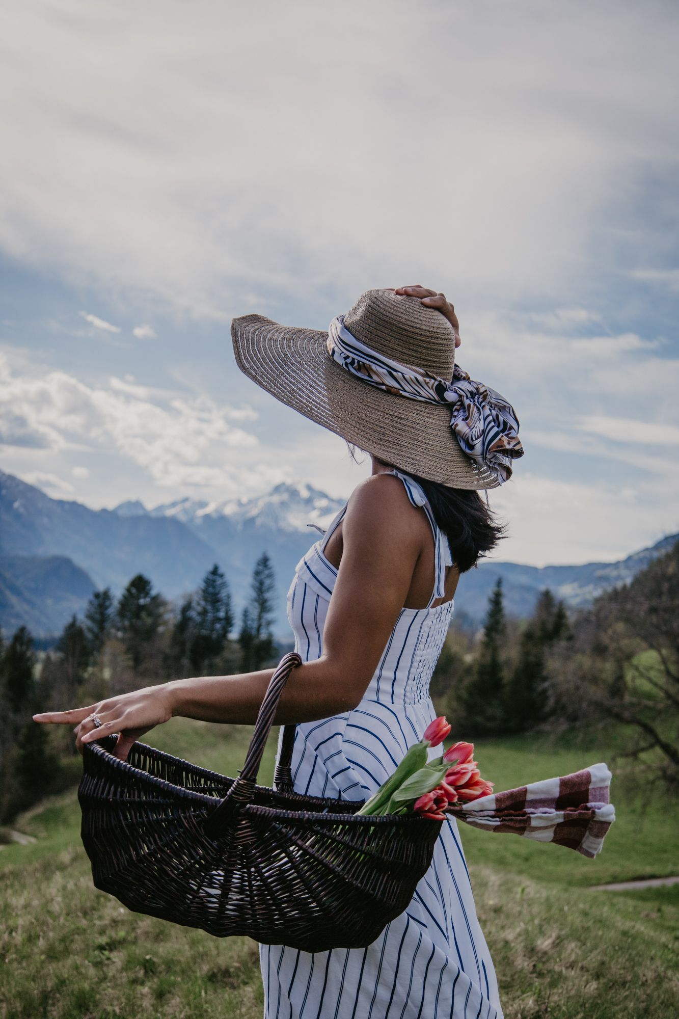 A person holding a dark wicker picnic basket against a backdrop of snow-capped mountains.