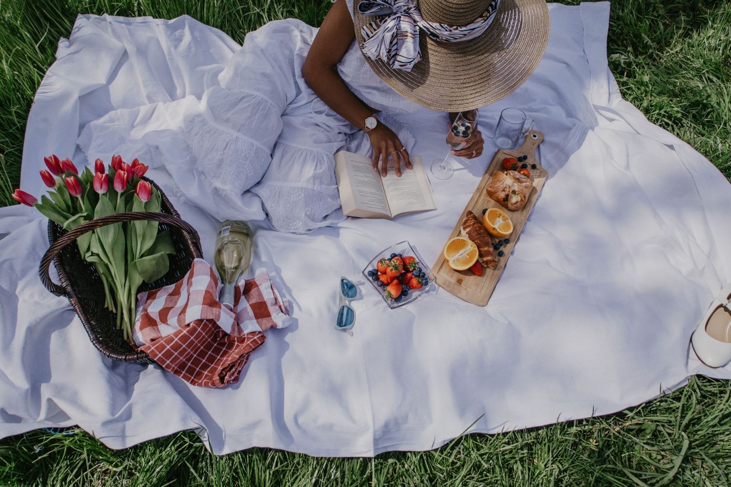 Picknick auf weißer Decke mit Obst, Gebäck, Blumen und einem Mädchen das ein Buch liest.