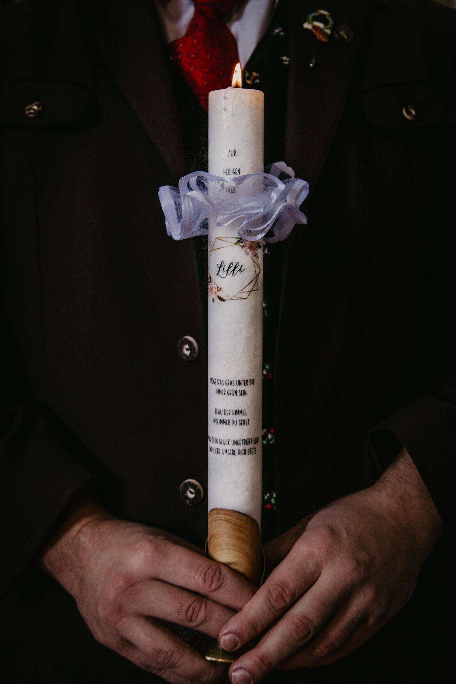 Man in a dark jacket holds a lit white baptism candle with a personalized label and white tulle ruffle.