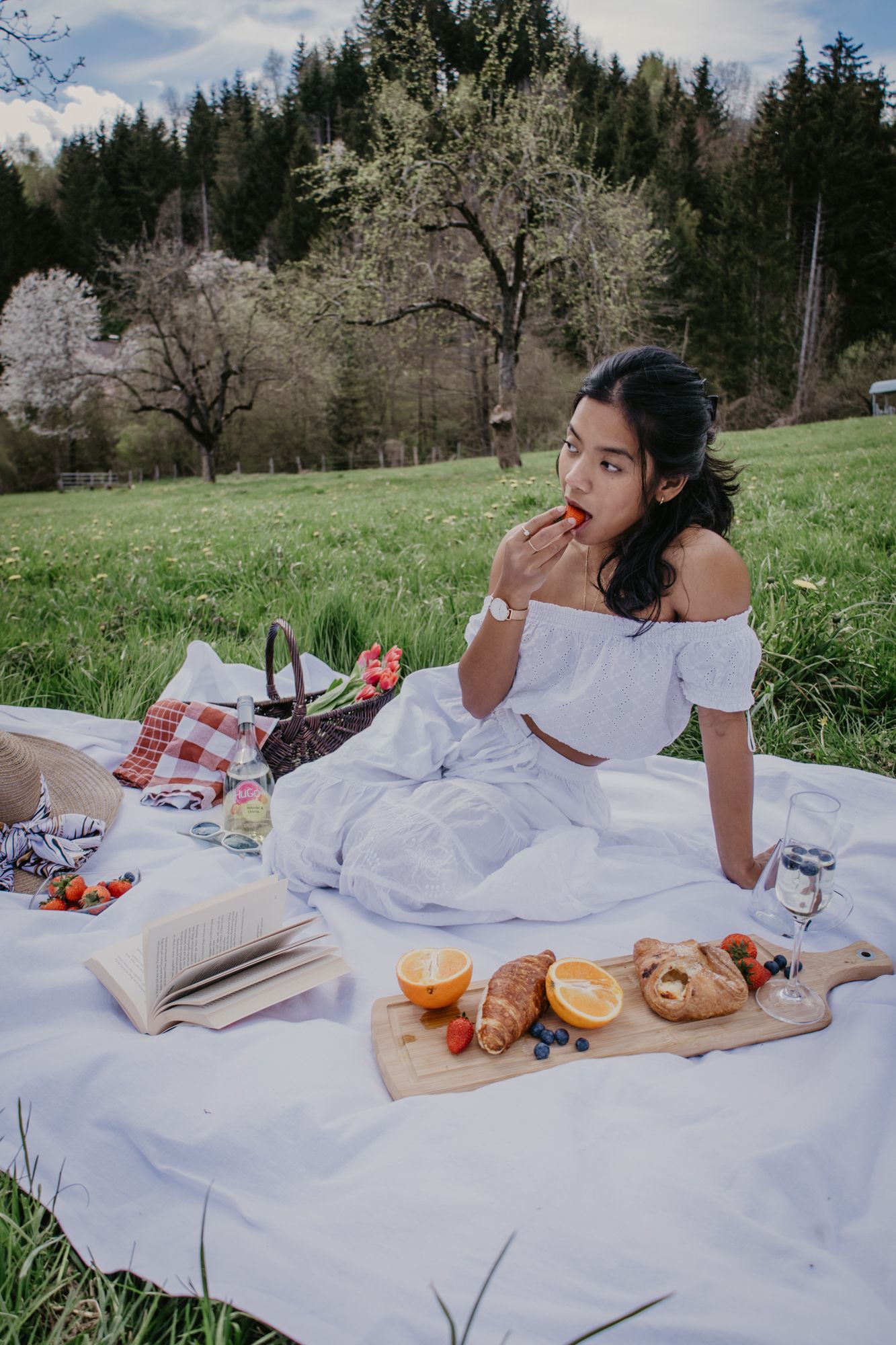 A serene picnic setup with fresh fruit, bread, and reading materials on a white blanket in nature.