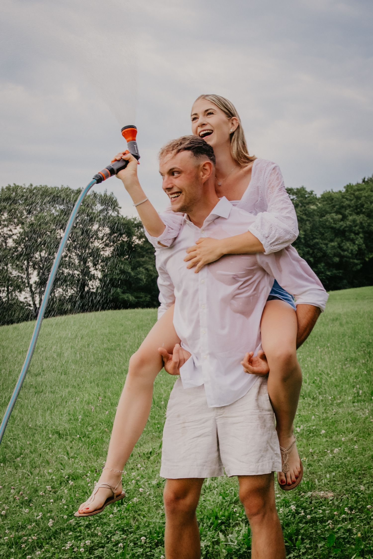 Happy couple playing with garden hose outdoors, woman riding piggyback on man's back laughing on green lawn.