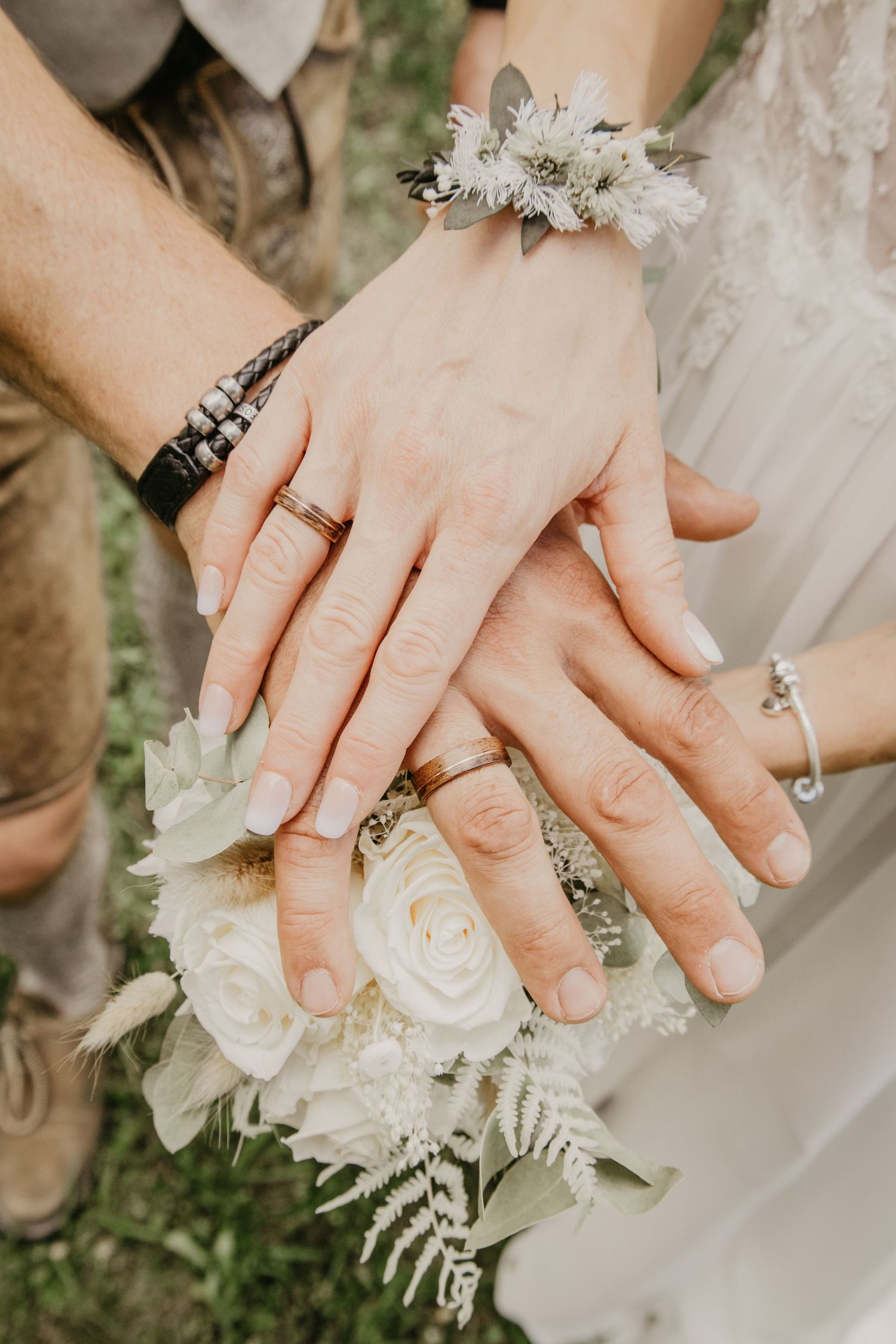Bride and groom hands stacked showing wedding rings on white rose bouquet with floral wrist corsage.