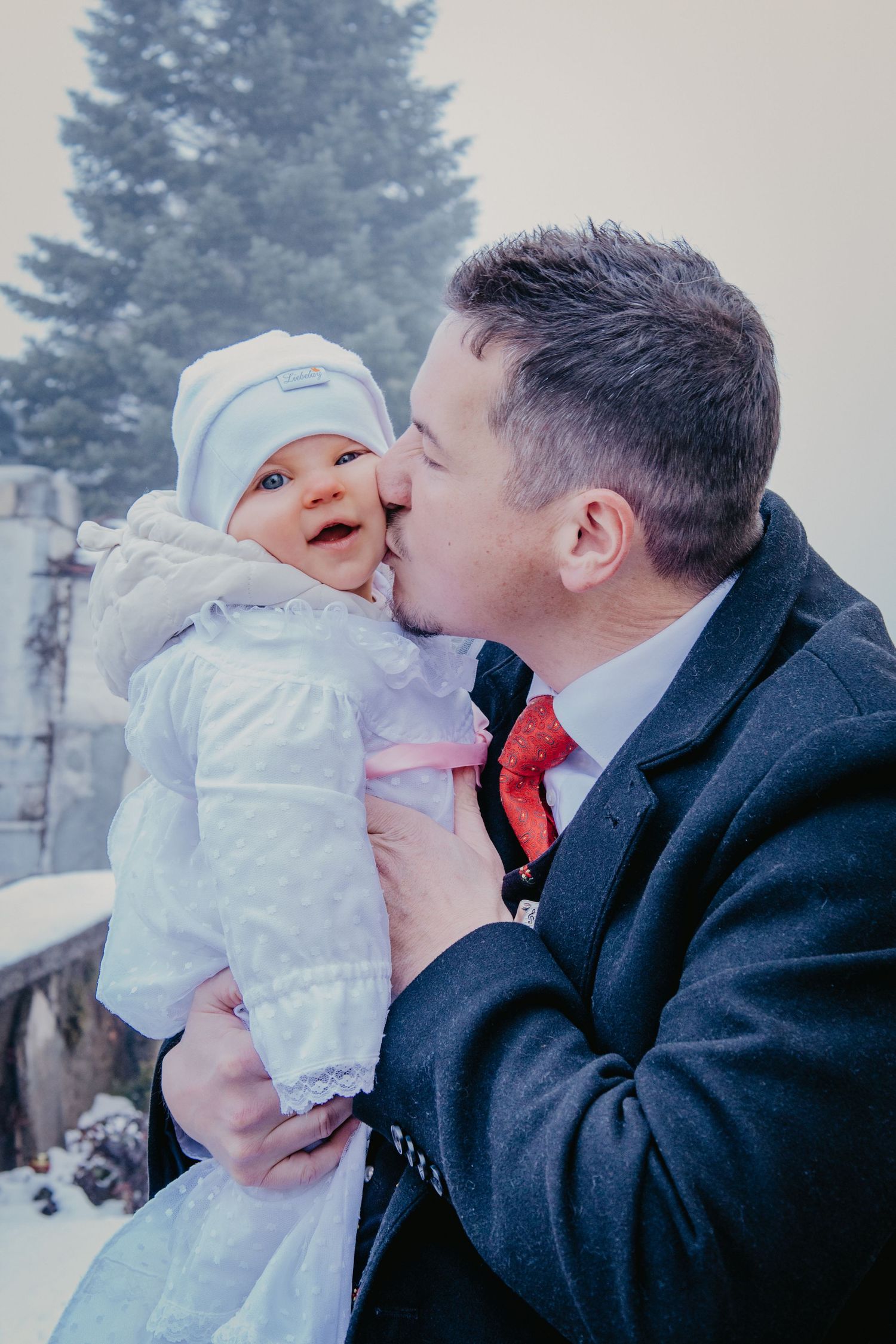 A man in a dark coat and red tie kisses the cheek of a smiling baby in a white outfit and bonnet outdoors in the snow.