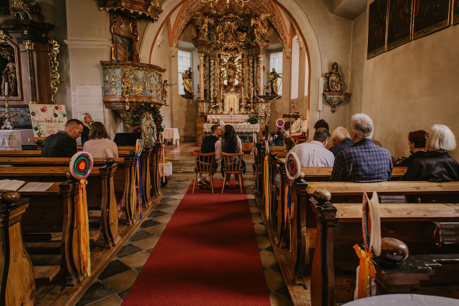 Worshippers sit in wooden pews along a red carpet leading to an ornate baroque altar inside a historic church.