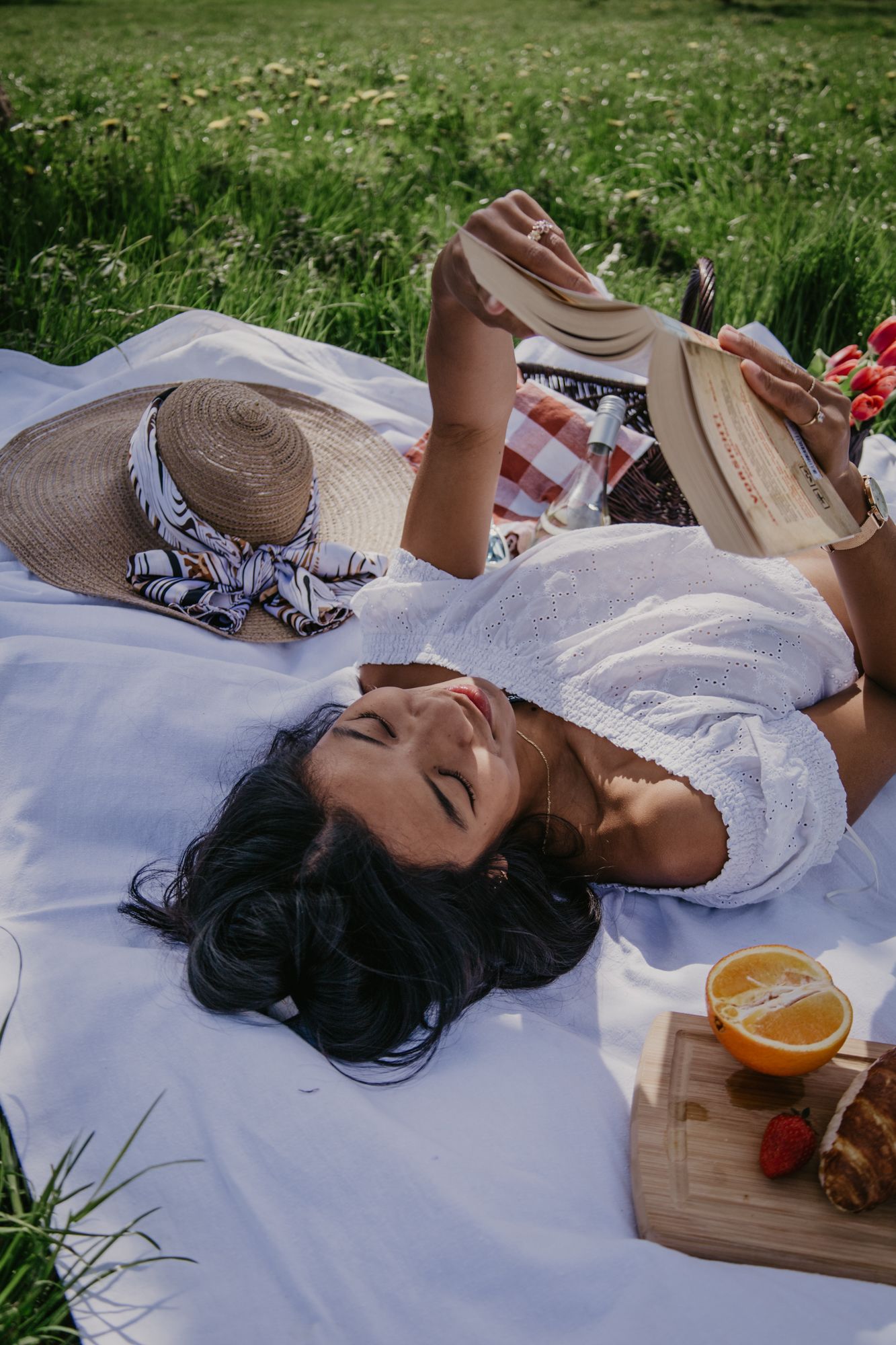 A peaceful picnic scene with someone relaxing on a white blanket in the grass with snacks and drinks.
