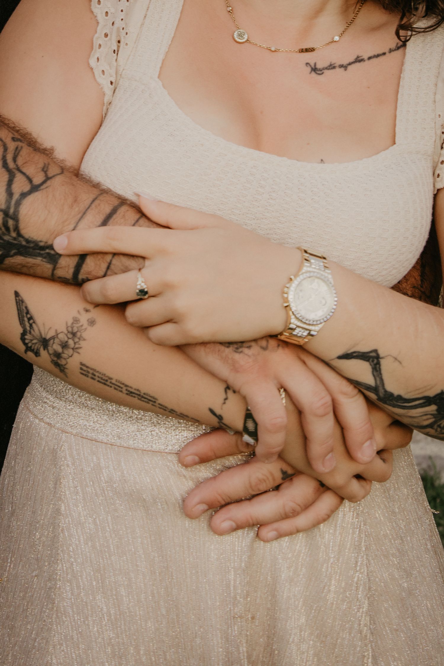 Close-up details of hands with tattoos and jewelry against a light-colored dress.