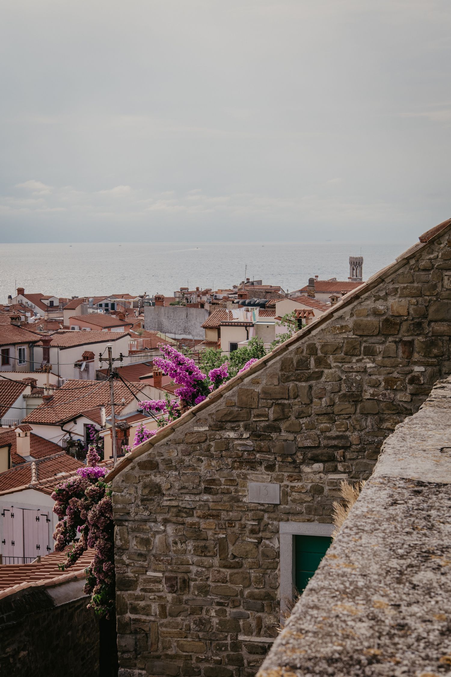 Ancient stone wall overlooks a Mediterranean town with red roofs and blooming bougainvillea.