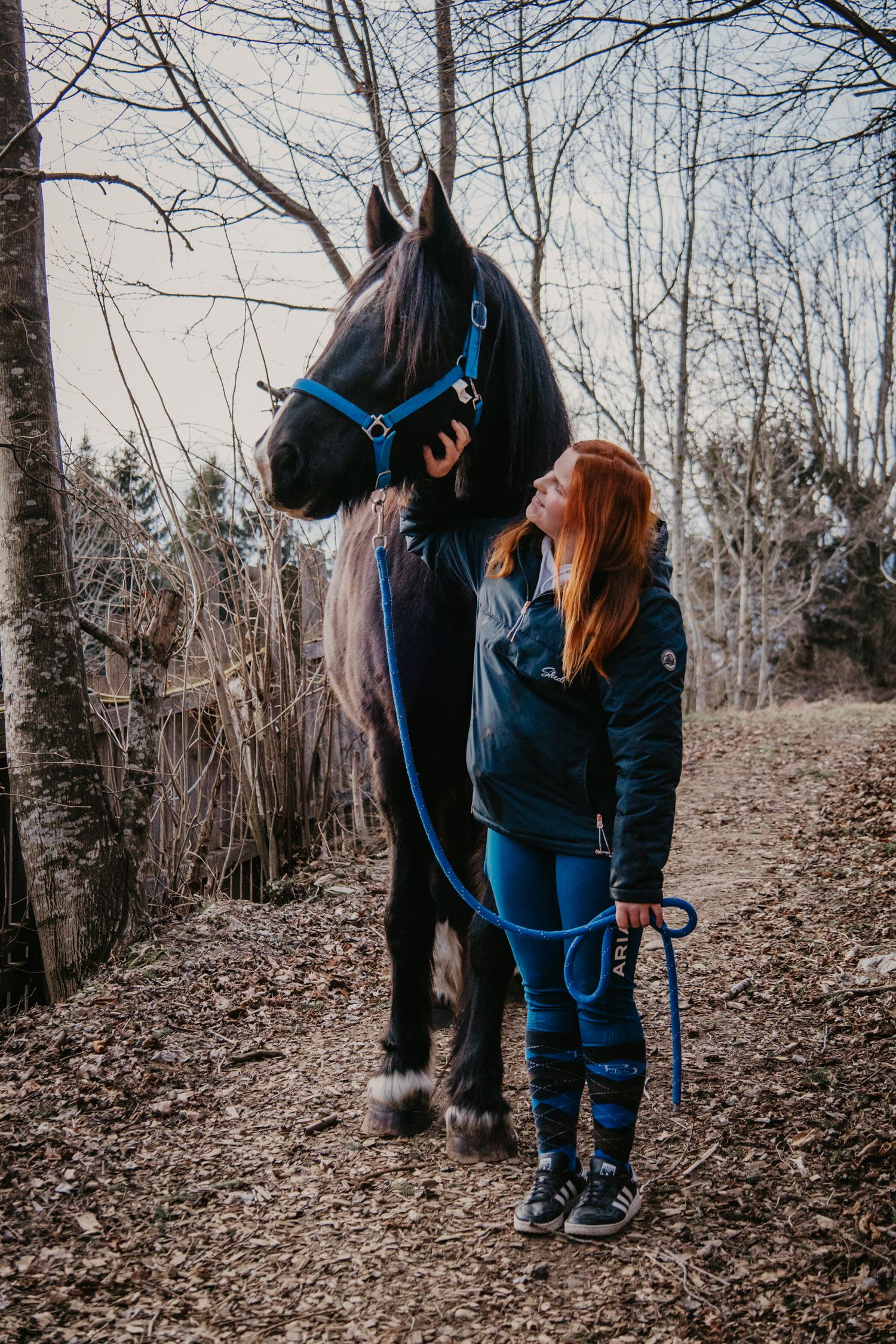 Person in dark clothing stands with black horse on autumn forest trail.