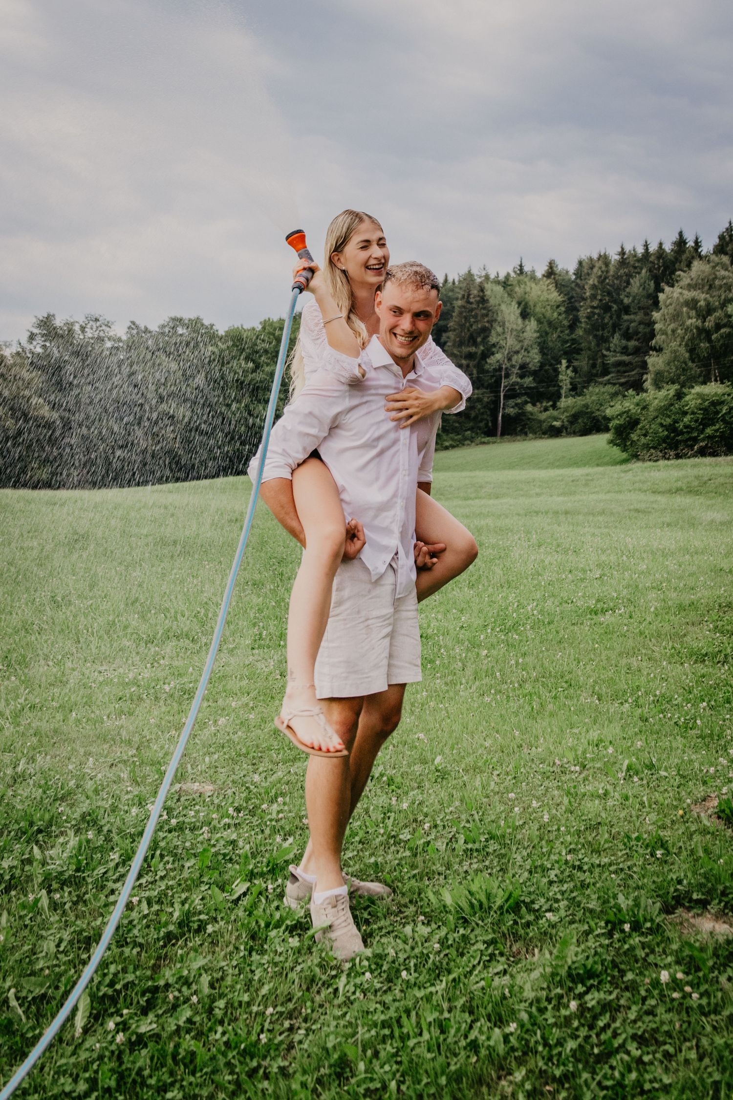 A happy couple plays with a garden hose on a green meadow, the man giving the woman a piggyback ride.
