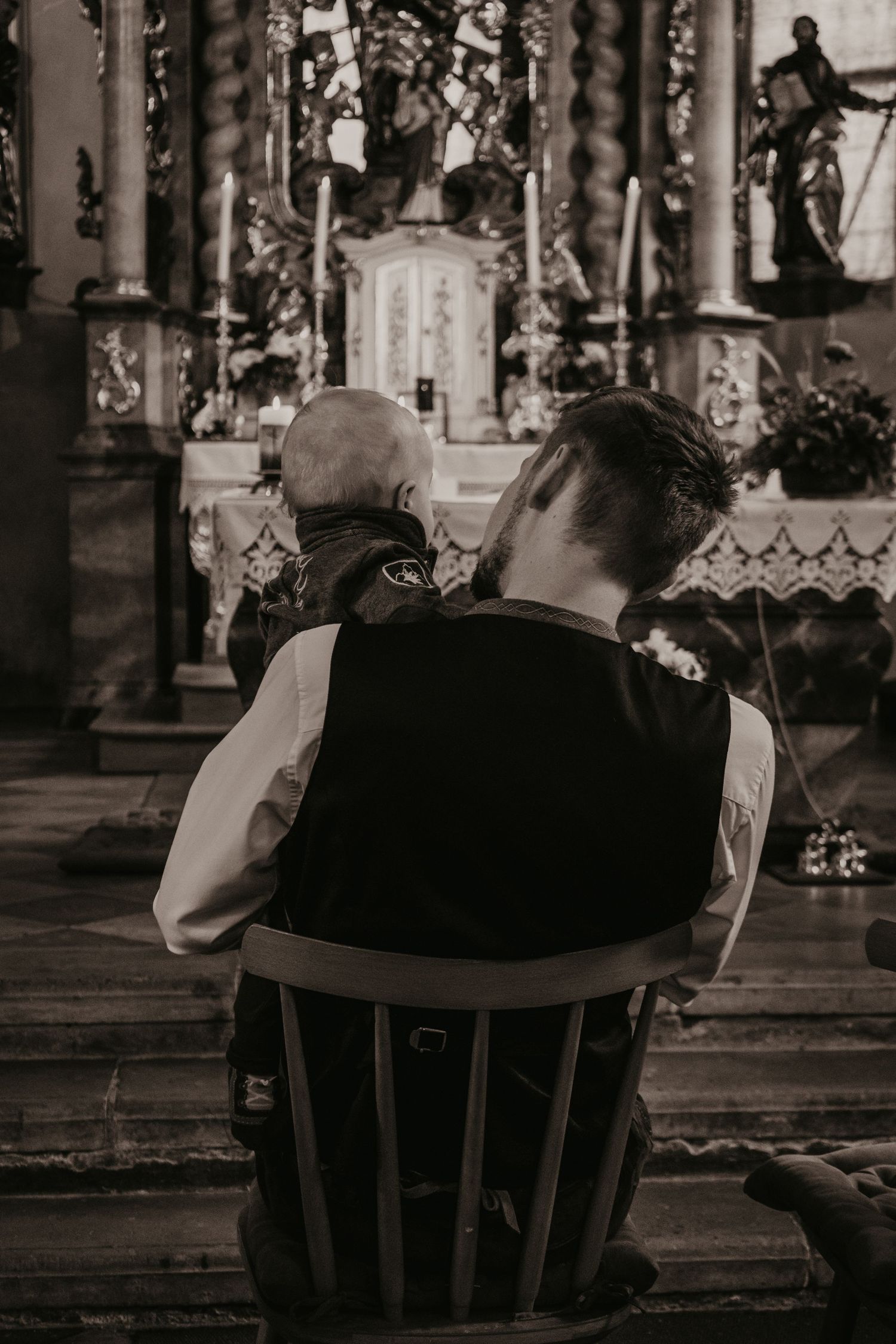 Man in a vest sits on a wooden chair holding a baby in front of an ornate church altar with candles, in black and white.