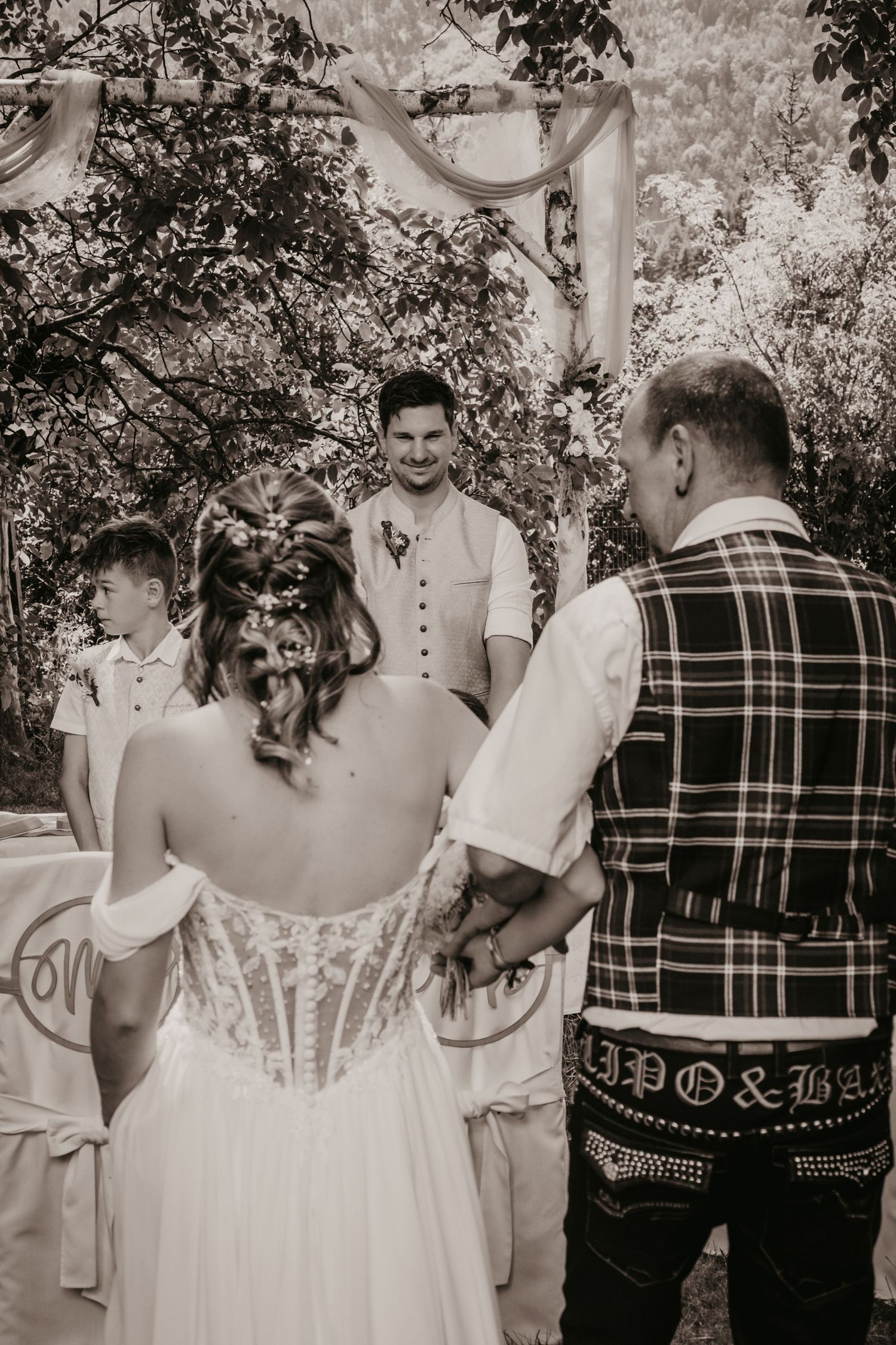Black and white wedding ceremony photo showing bride in corset gown with groom and officiant outdoors under floral arch.