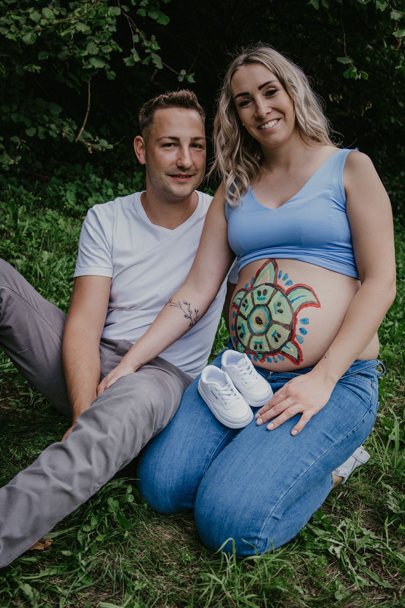 Couple sitting on grass during maternity shoot with baby shoes and painted belly art.
