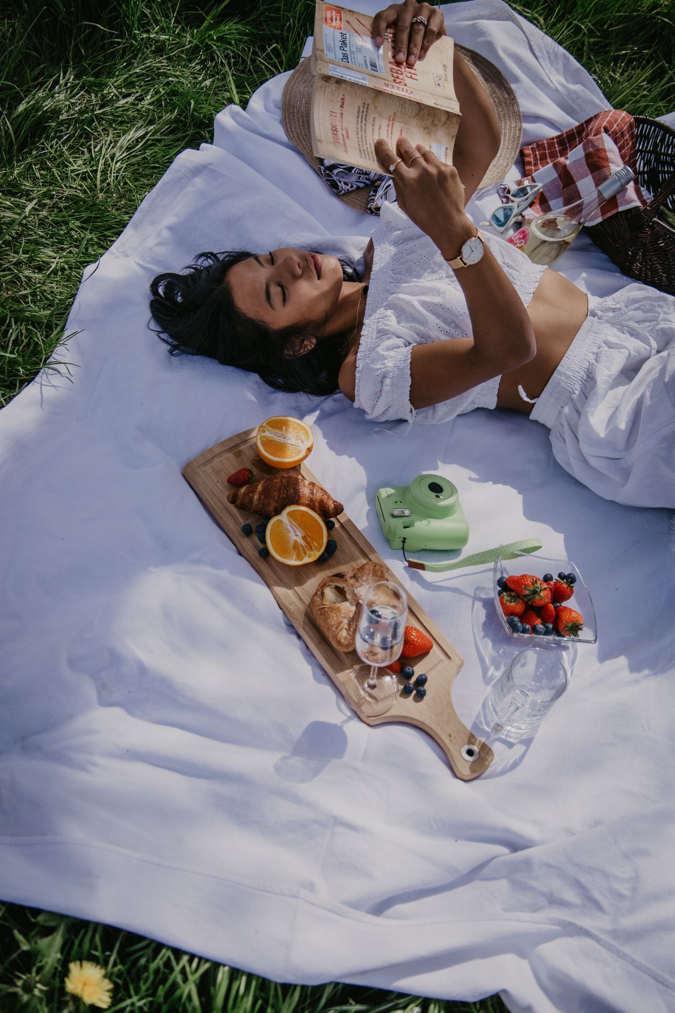 A cozy outdoor picnic featuring books, citrus fruits, and snacks on a white blanket in the grass.