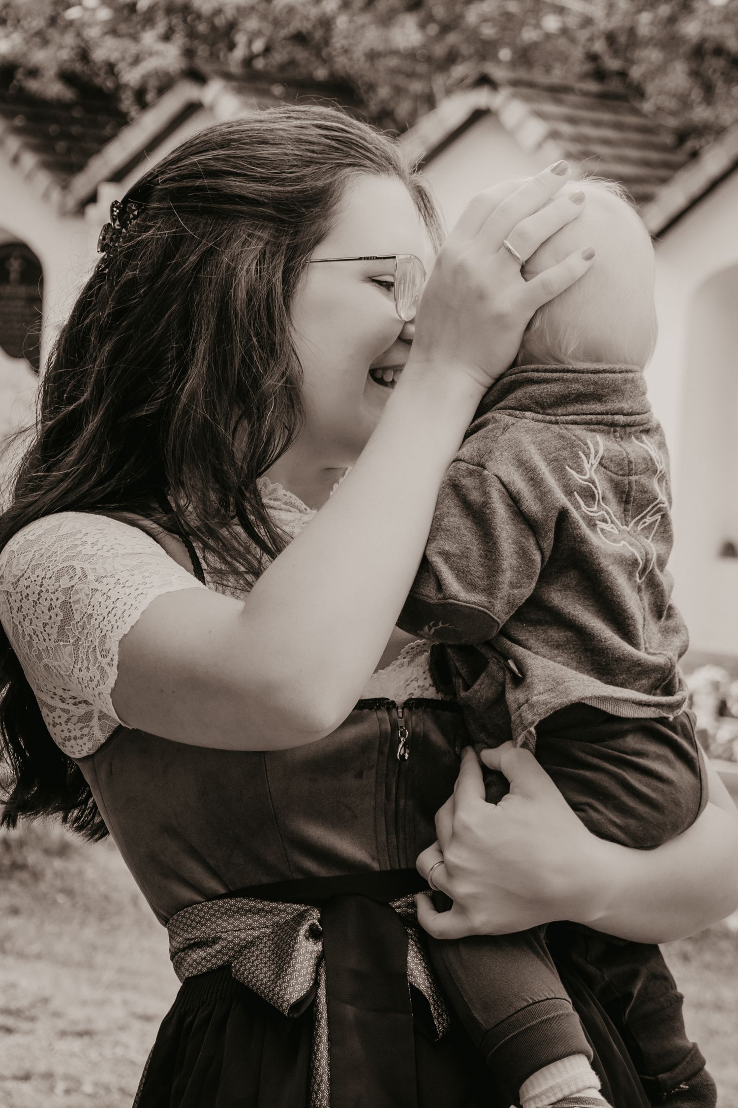 Smiling woman with glasses and long hair lovingly holds a toddler against her cheek in a black and white outdoor portrait.