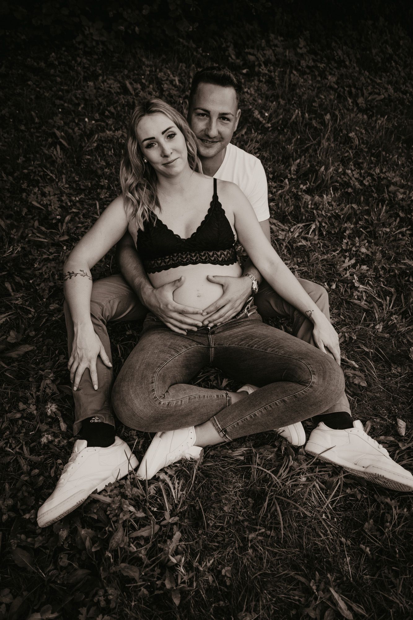 A black and white maternity photo of a couple sitting together outdoors.