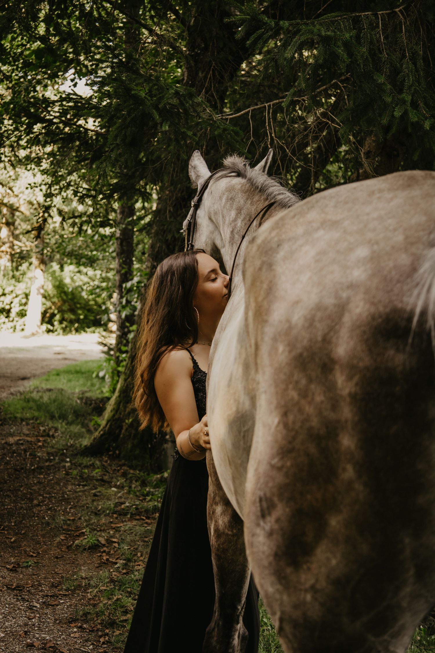 A tender moment captured between a grey horse and someone in a black dress in a wooded setting.