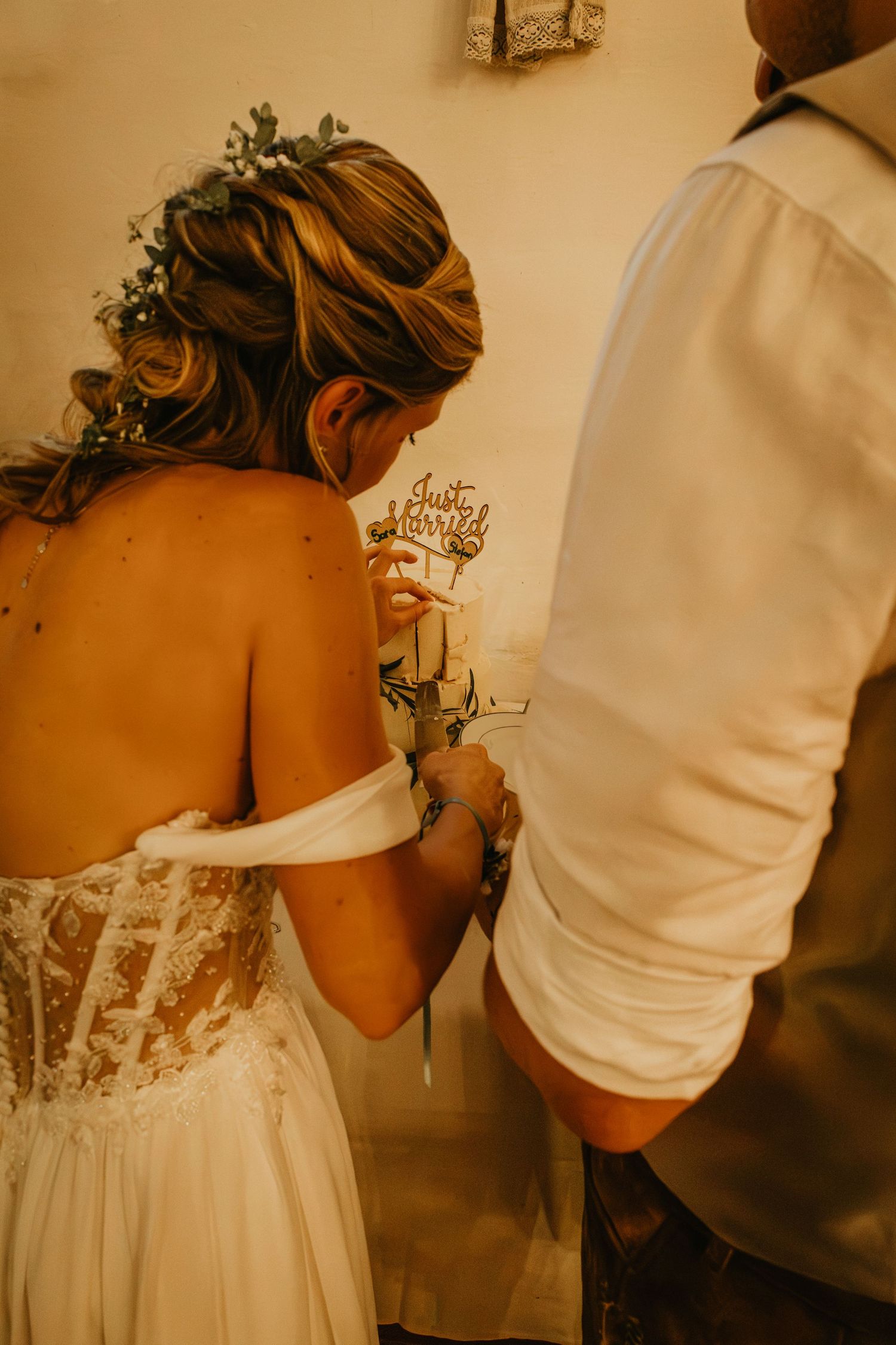 Bride in lace backless wedding dress with floral hair piece cuts cake with groom holding Just Married topper.