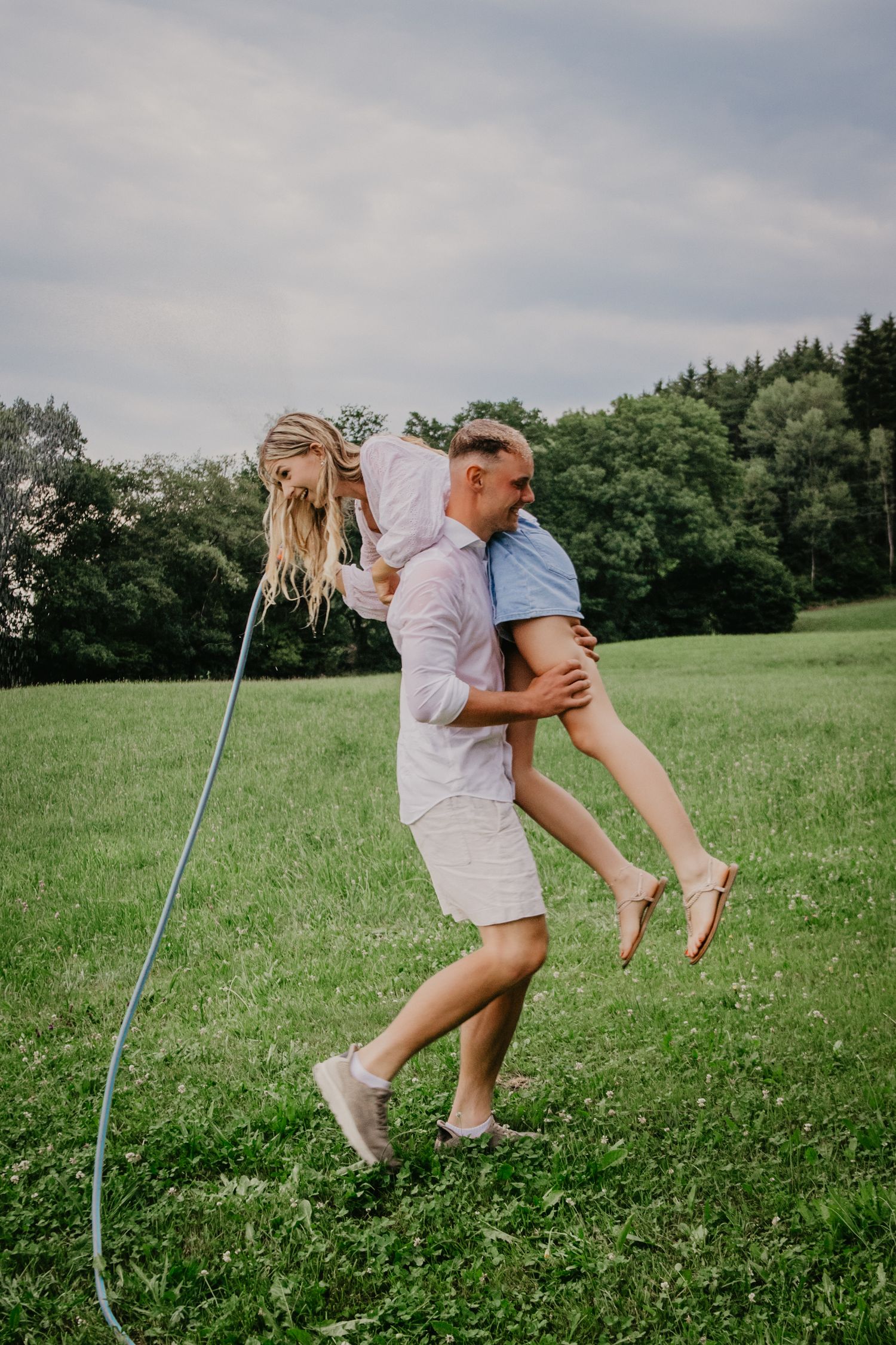Man carrying woman over his shoulder in a green field, both laughing and enjoying a playful outdoor moment.