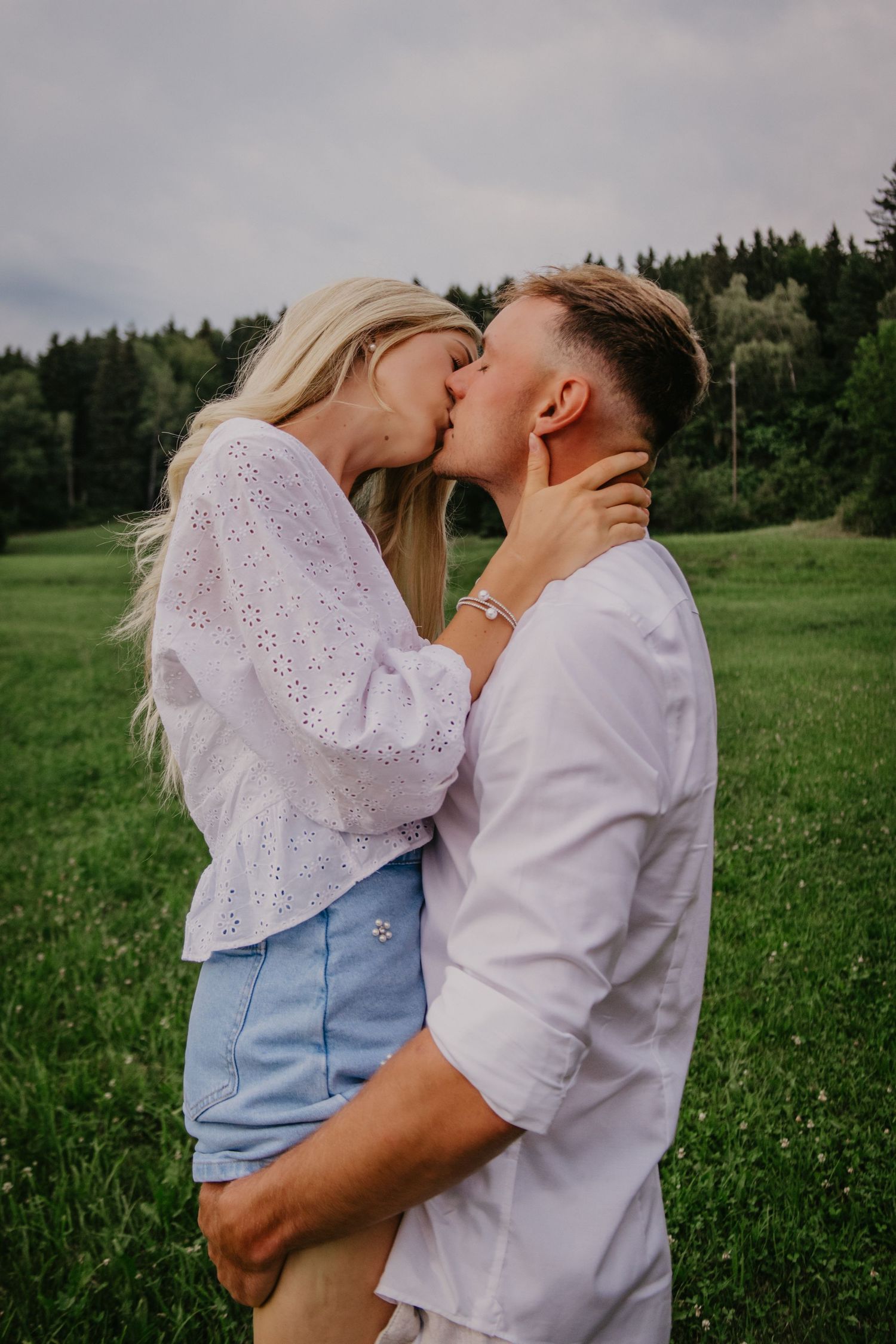 A couple shares a romantic kiss in a lush green meadow, the woman in a white blouse and blue shorts, the man in a white shirt.