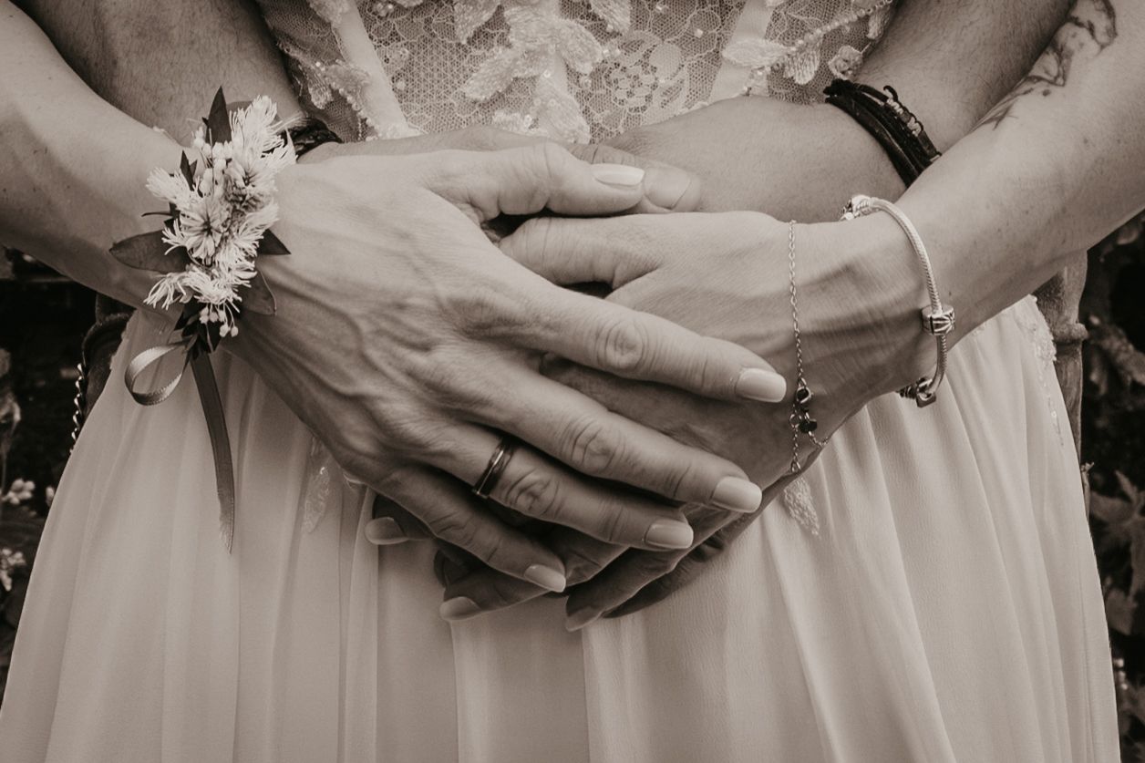 Close-up black and white photo of bride and groom hands clasped together, showing wedding rings and floral wrist corsage.