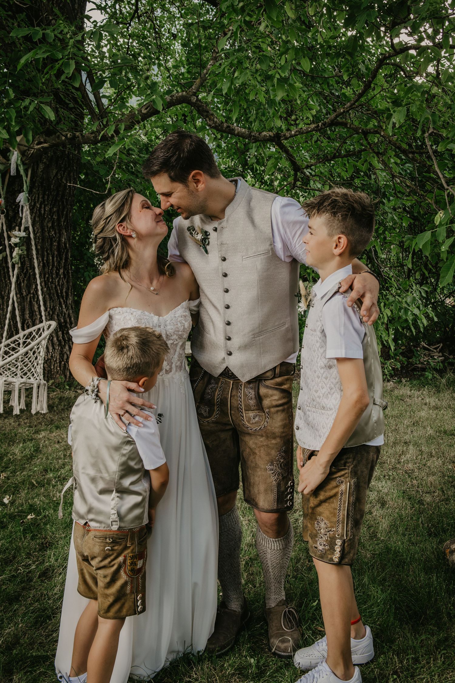 Bride and groom in traditional Bavarian attire share a romantic moment with two young boys in a lush garden setting.