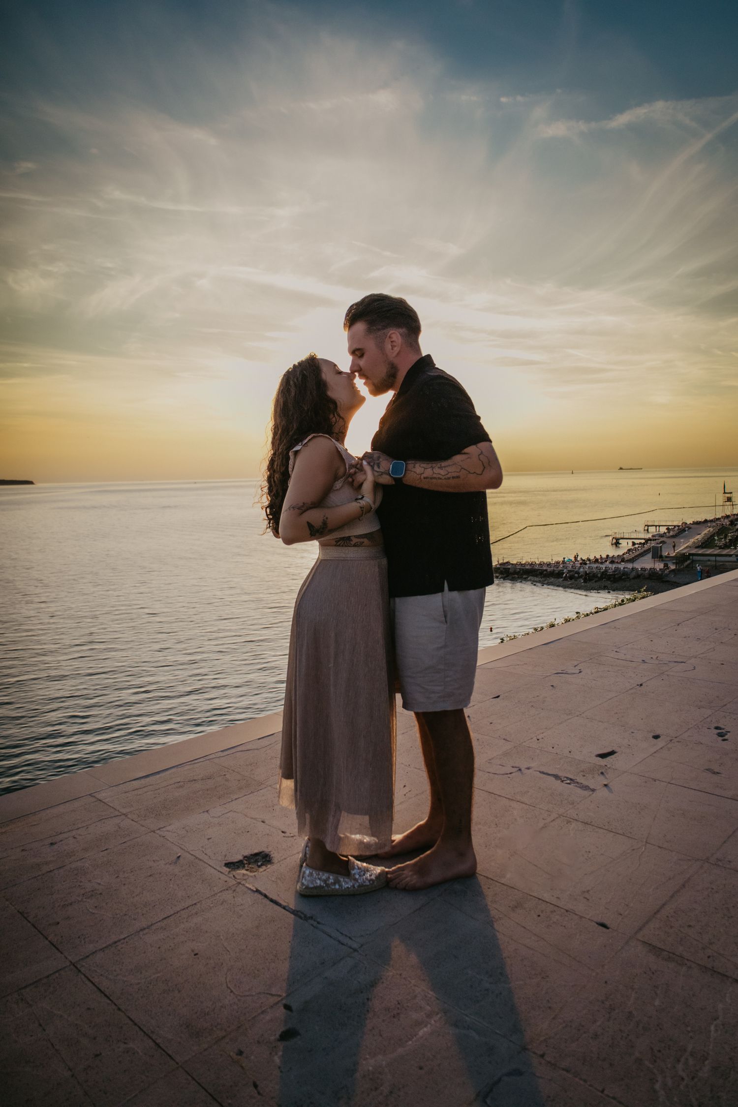 A couple shares an intimate moment at sunset on a concrete pier overlooking a calm ocean.