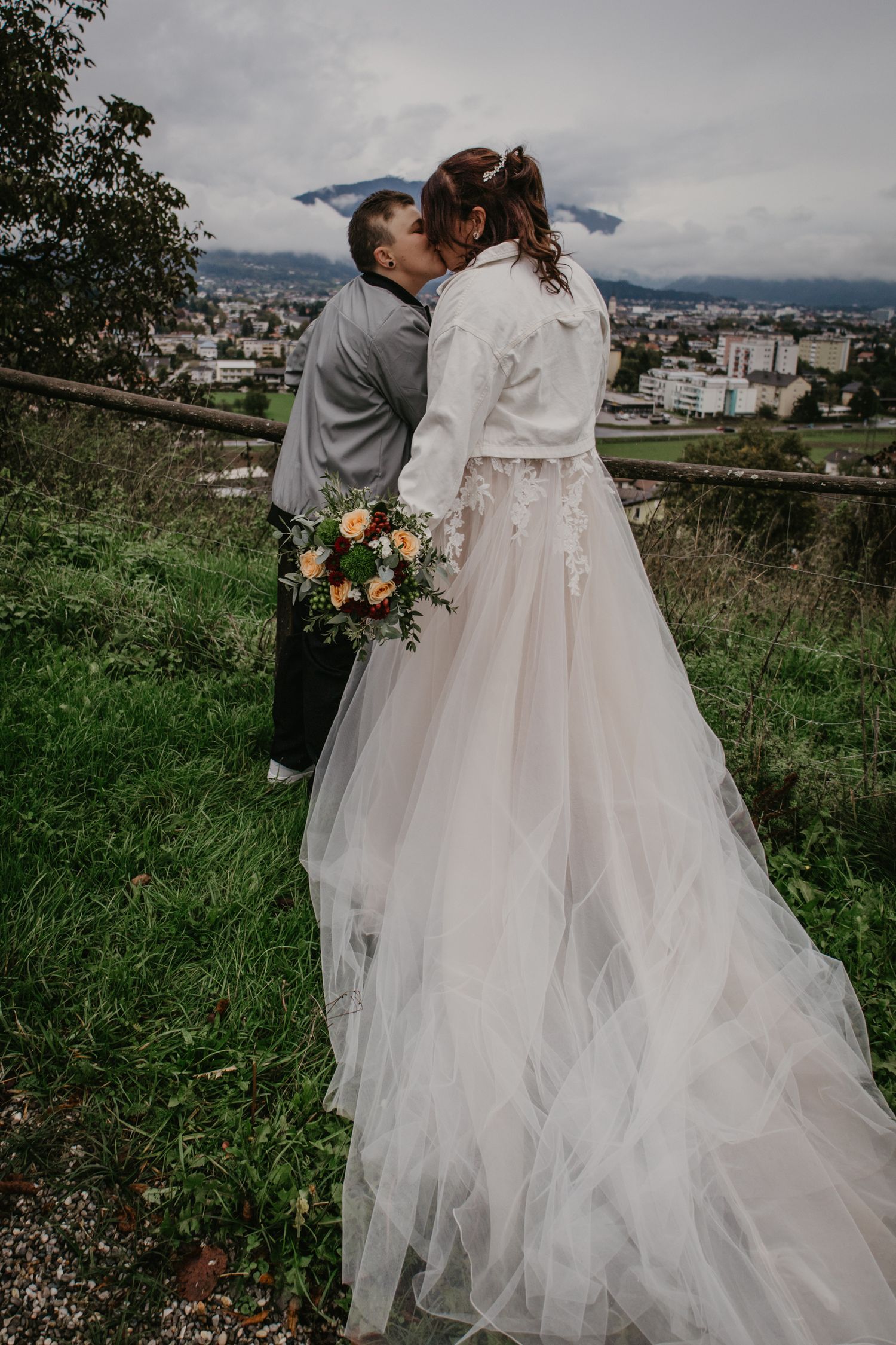 Couple in wedding attire walking on hilltop overlooking city landscape.
