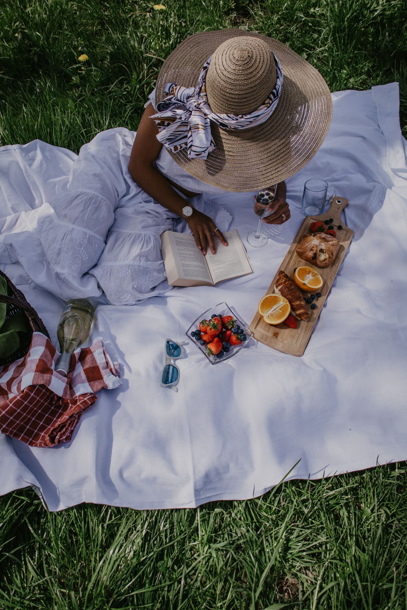 Overhead view of picnic setup with straw hat, food, and drinks on white blanket.