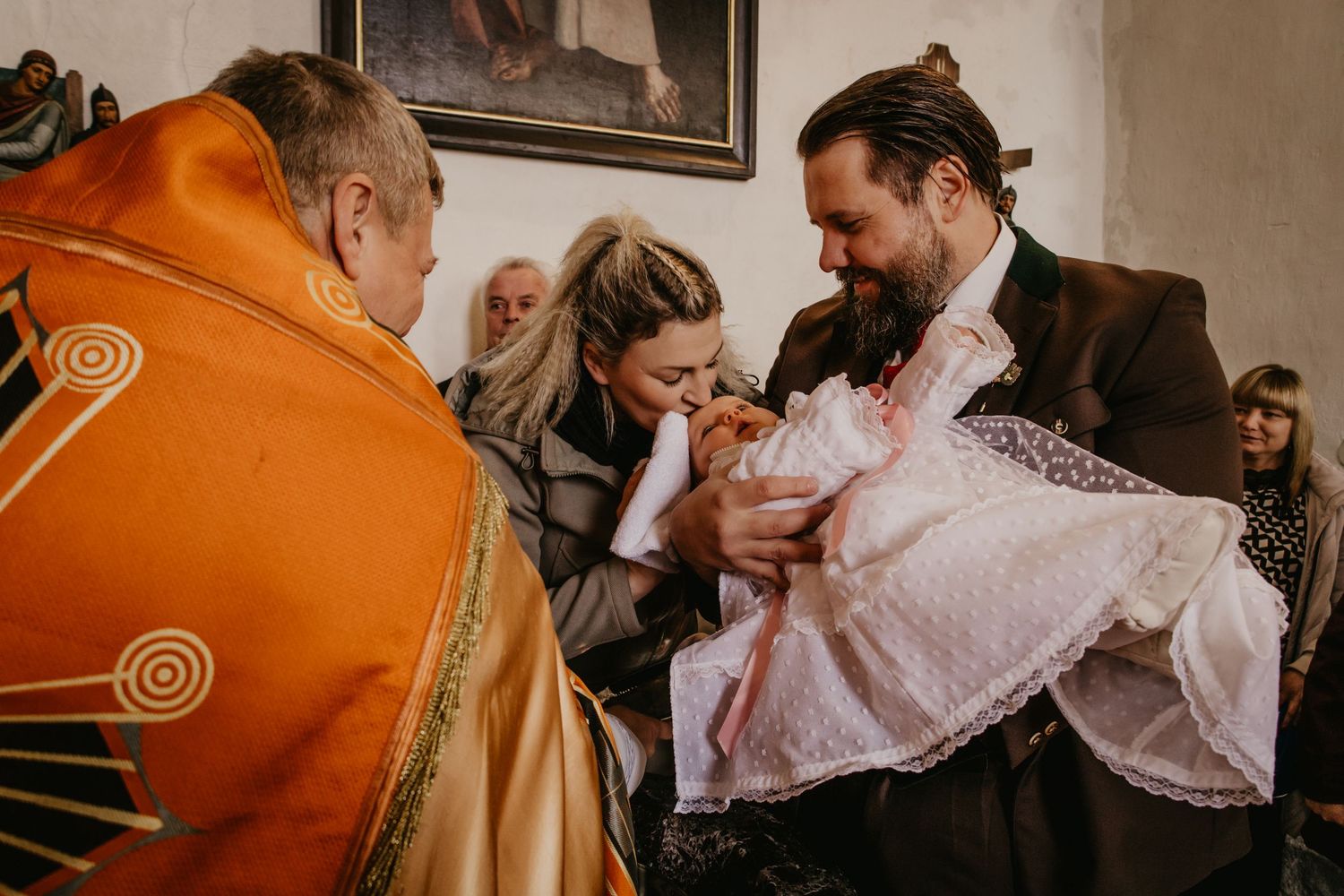 A bearded man in a dark coat holds a baby in a white lace christening gown beside a priest in orange vestments during a church baptism.