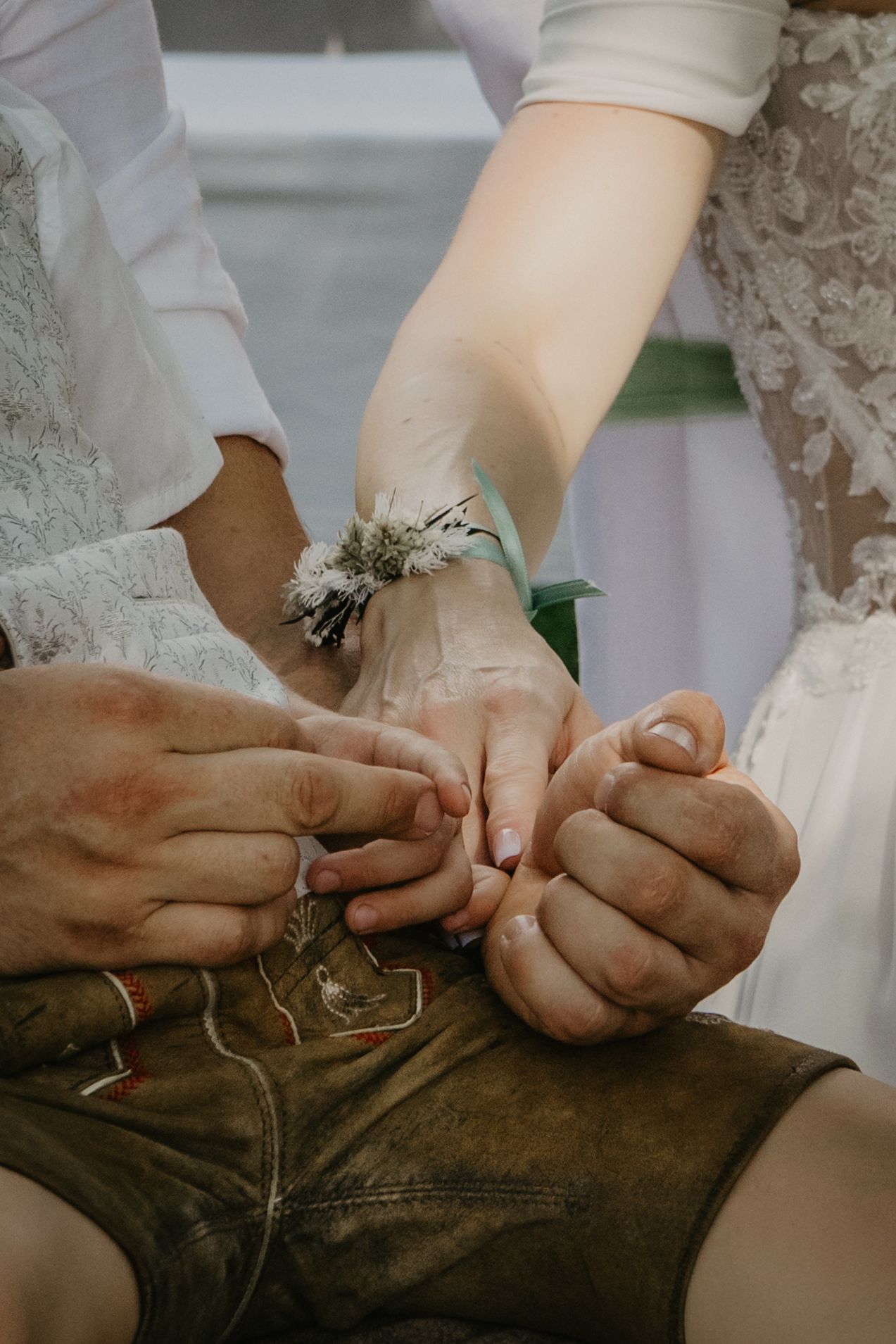 Close-up of hands holding together at a wedding ceremony, bride wearing floral wrist corsage and lace dress.