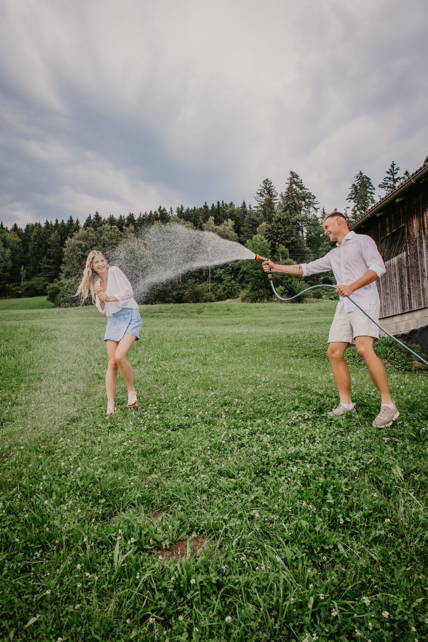 A man playfully sprays a laughing woman with a garden hose on a green meadow, with forest and barn in background.