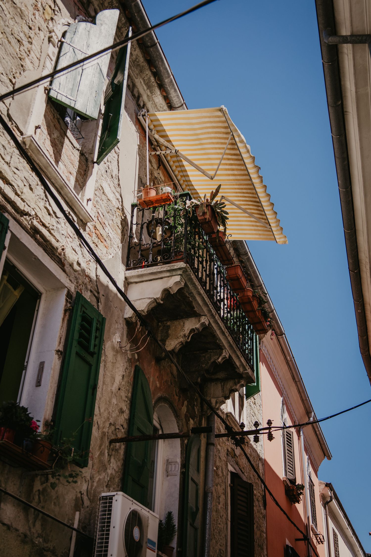 Historic stone buildings with ornate balconies and green shutters along a narrow alley.